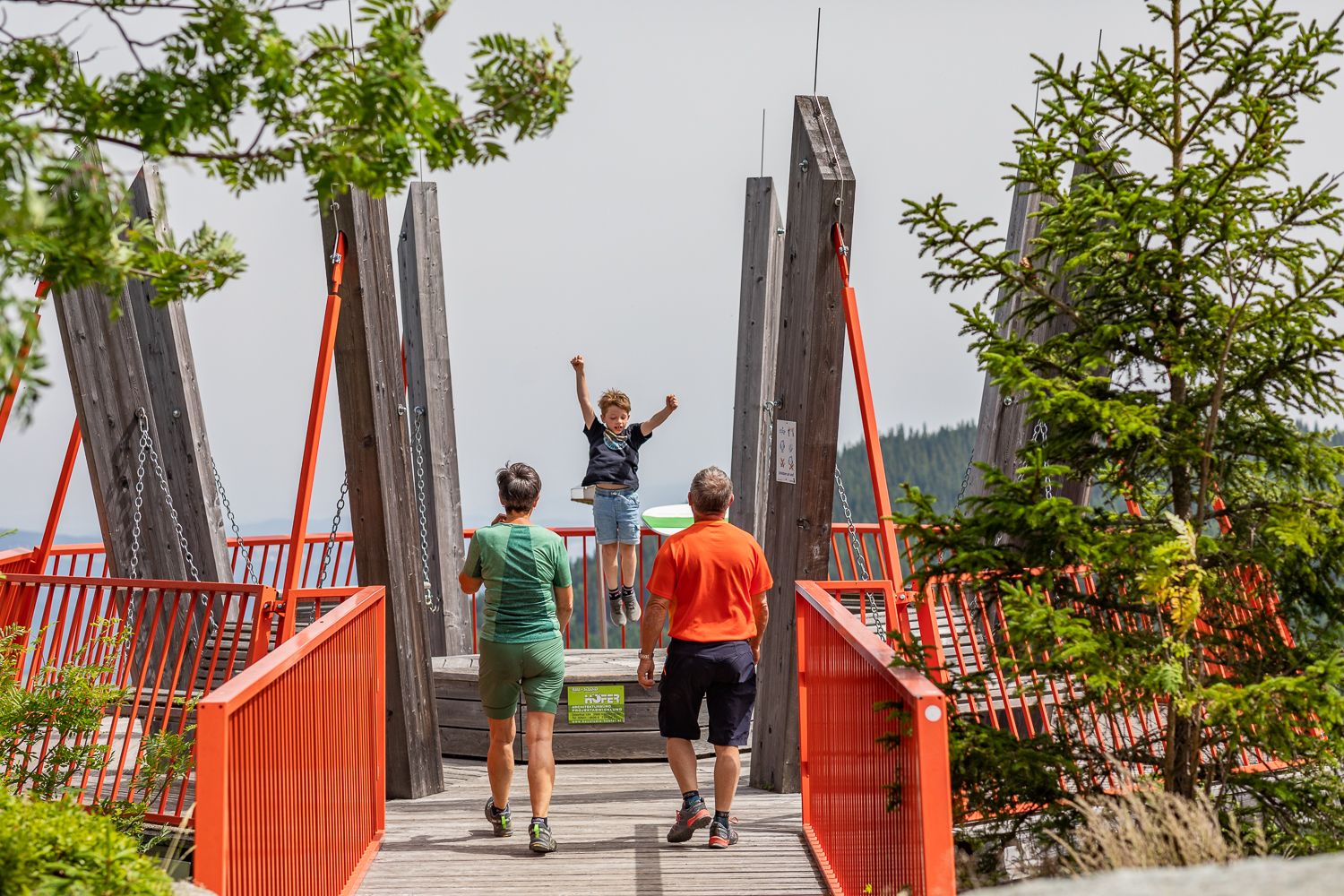 A child swings on a viewing platform with red railings while two adults look on.