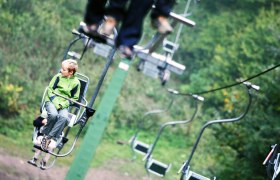 A boy in a green jacket sits on a chair lift, surrounded by green nature.
