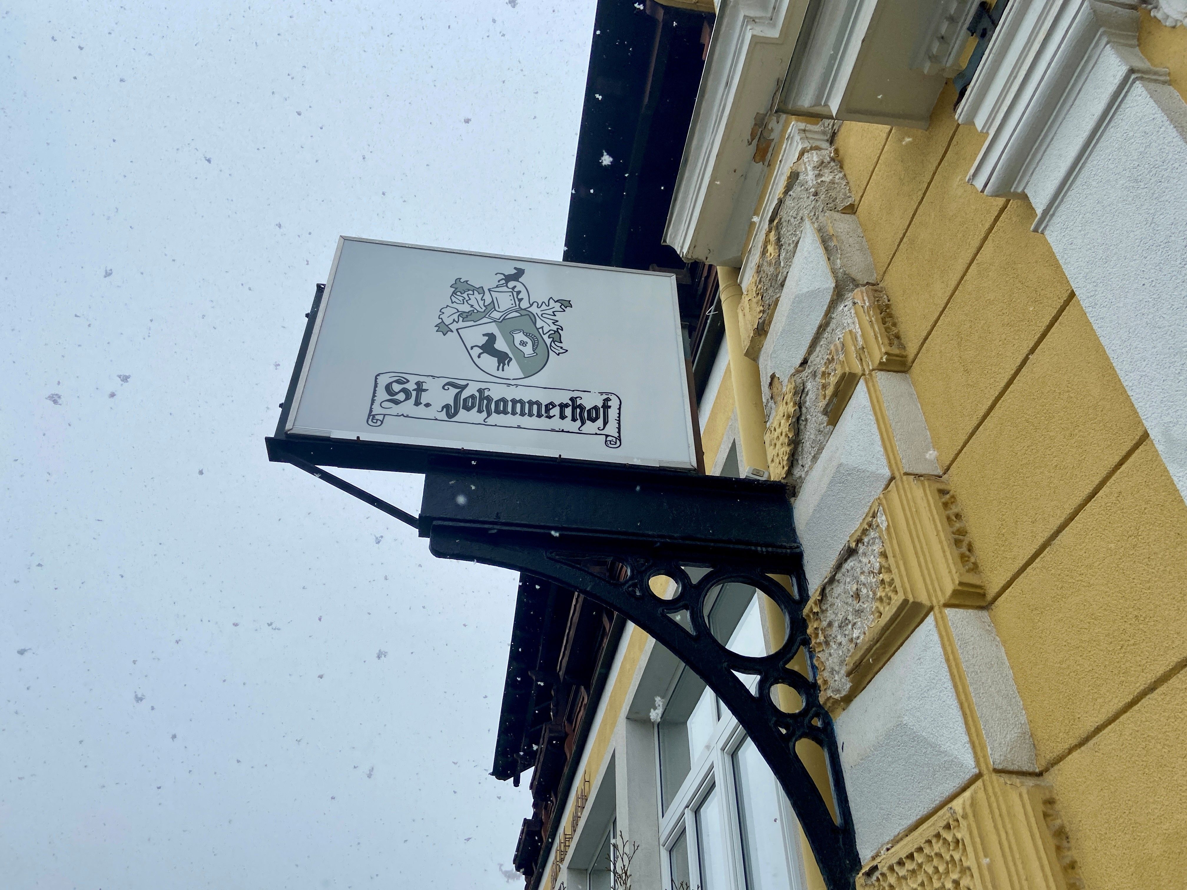 St. Johannerhof sign on a yellow building wall with snowflakes in the background.