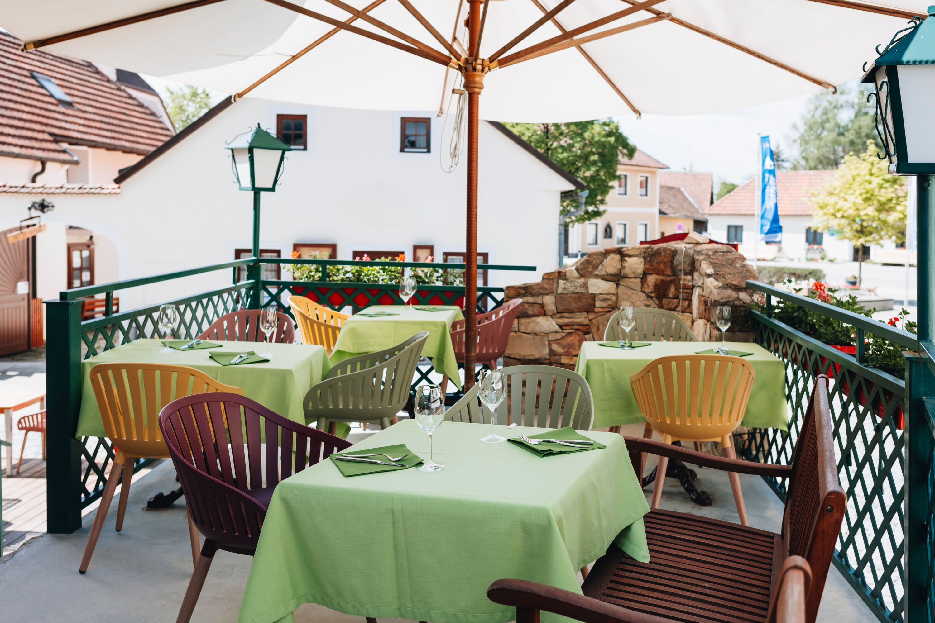 A guest garden with green tablecloths and colorful chairs under a parasol, surrounded by buildings and plants.