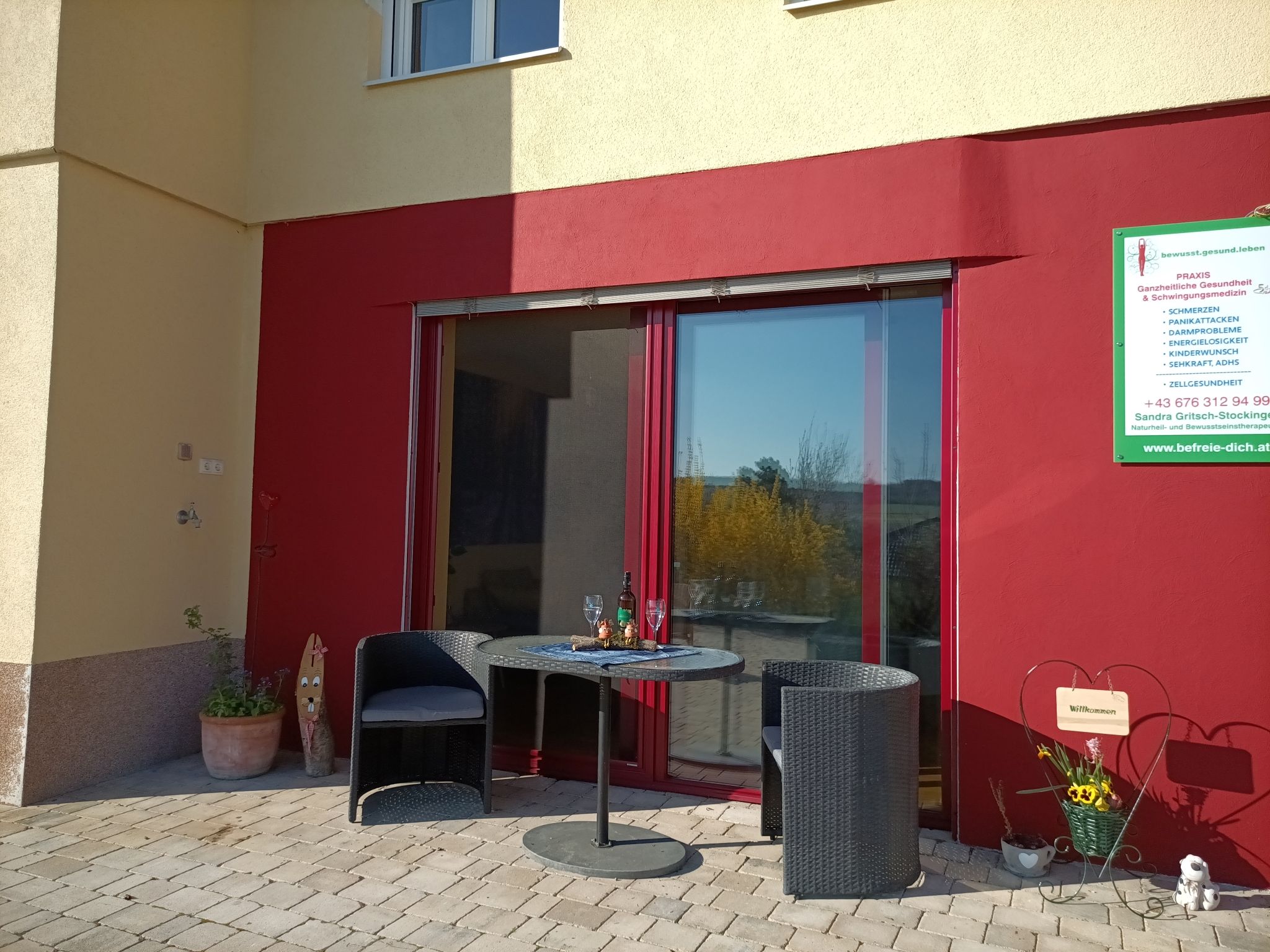 Terrace with two chairs and a table in front of a red wall, next to it a sign with information.