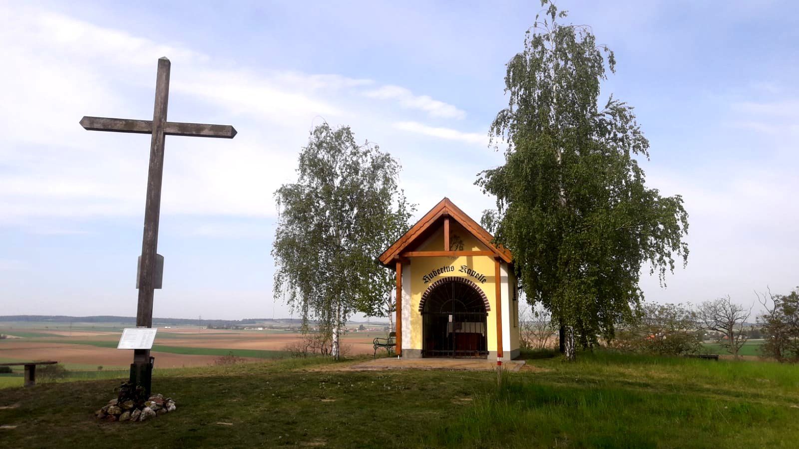 A wooden cross and a small chapel on a hill with trees and a wide view over the landscape.
