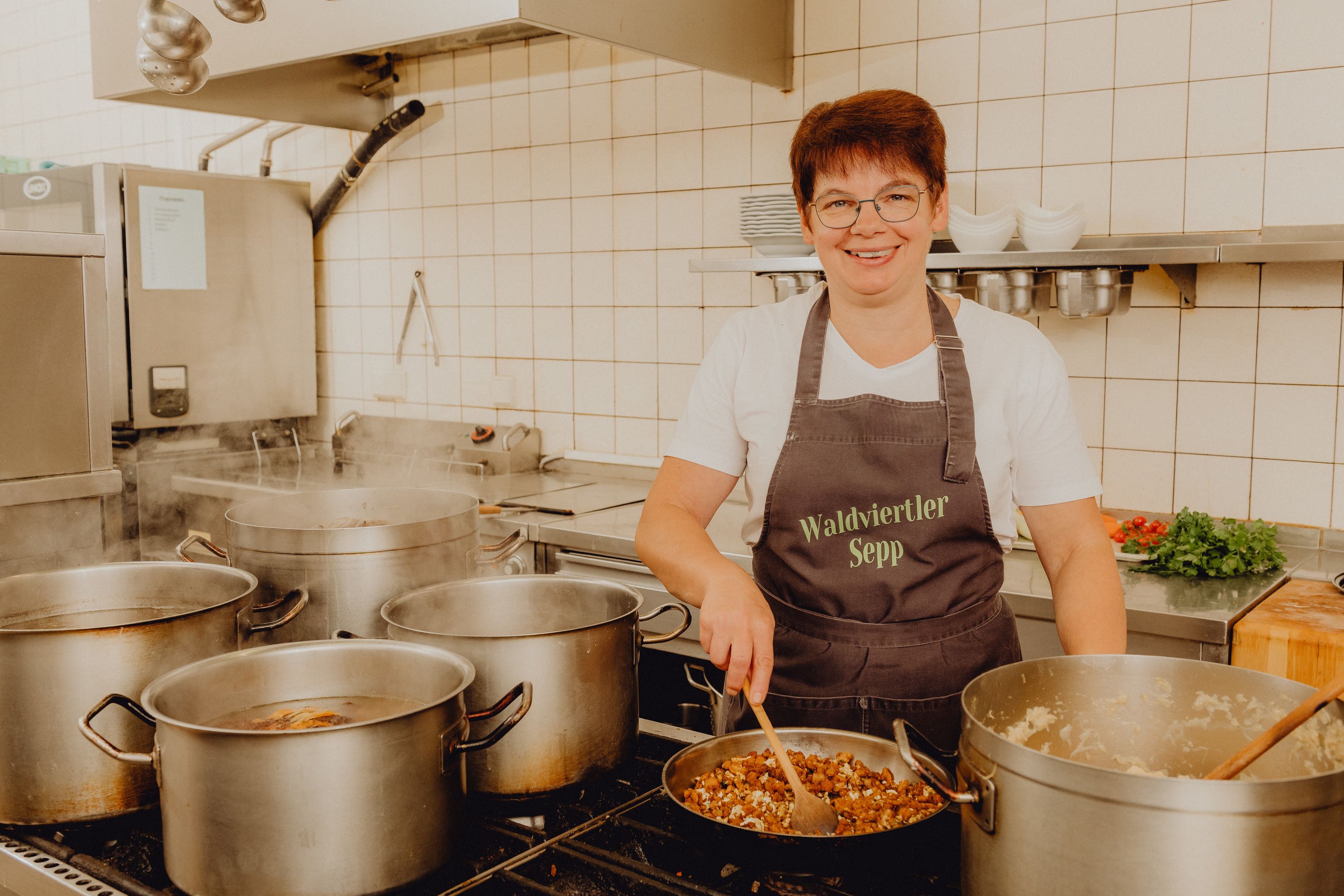 A cook in a professional kitchen serving several pots on a stove.