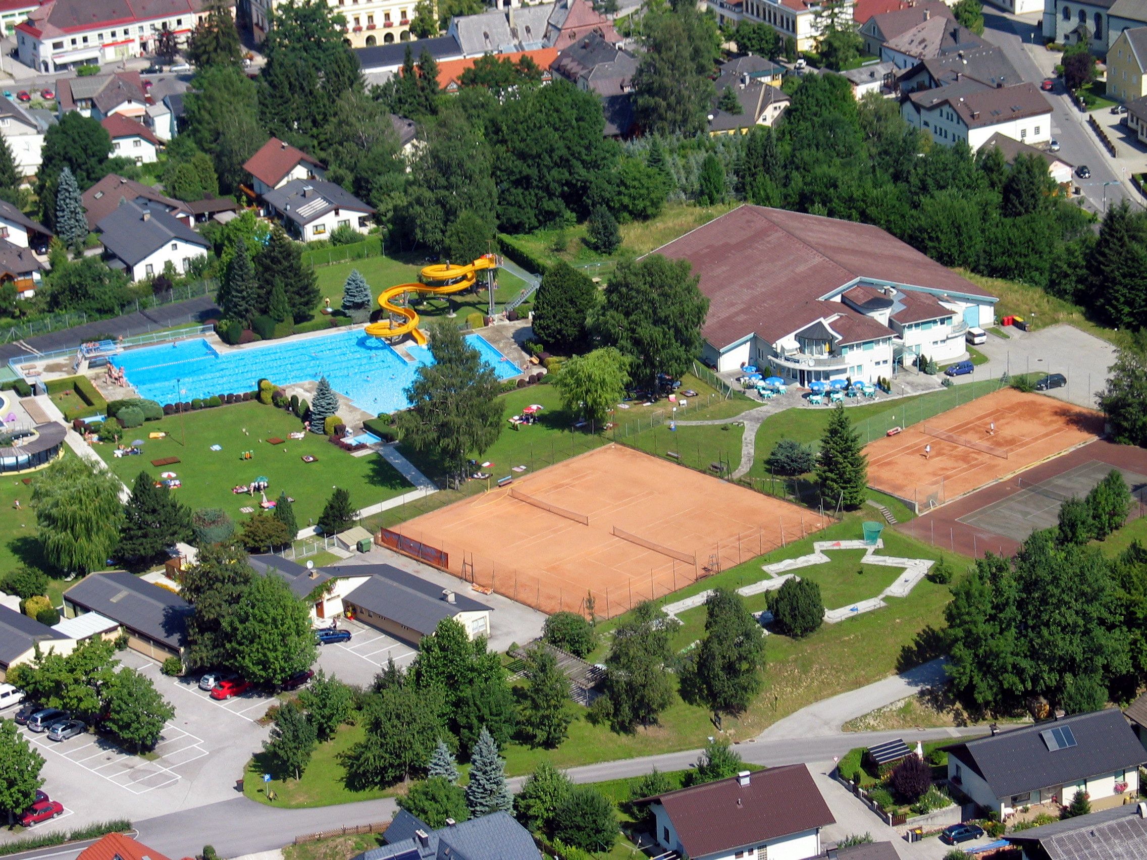 Aerial view of an outdoor pool with swimming pool, slide and tennis courts in a residential area.