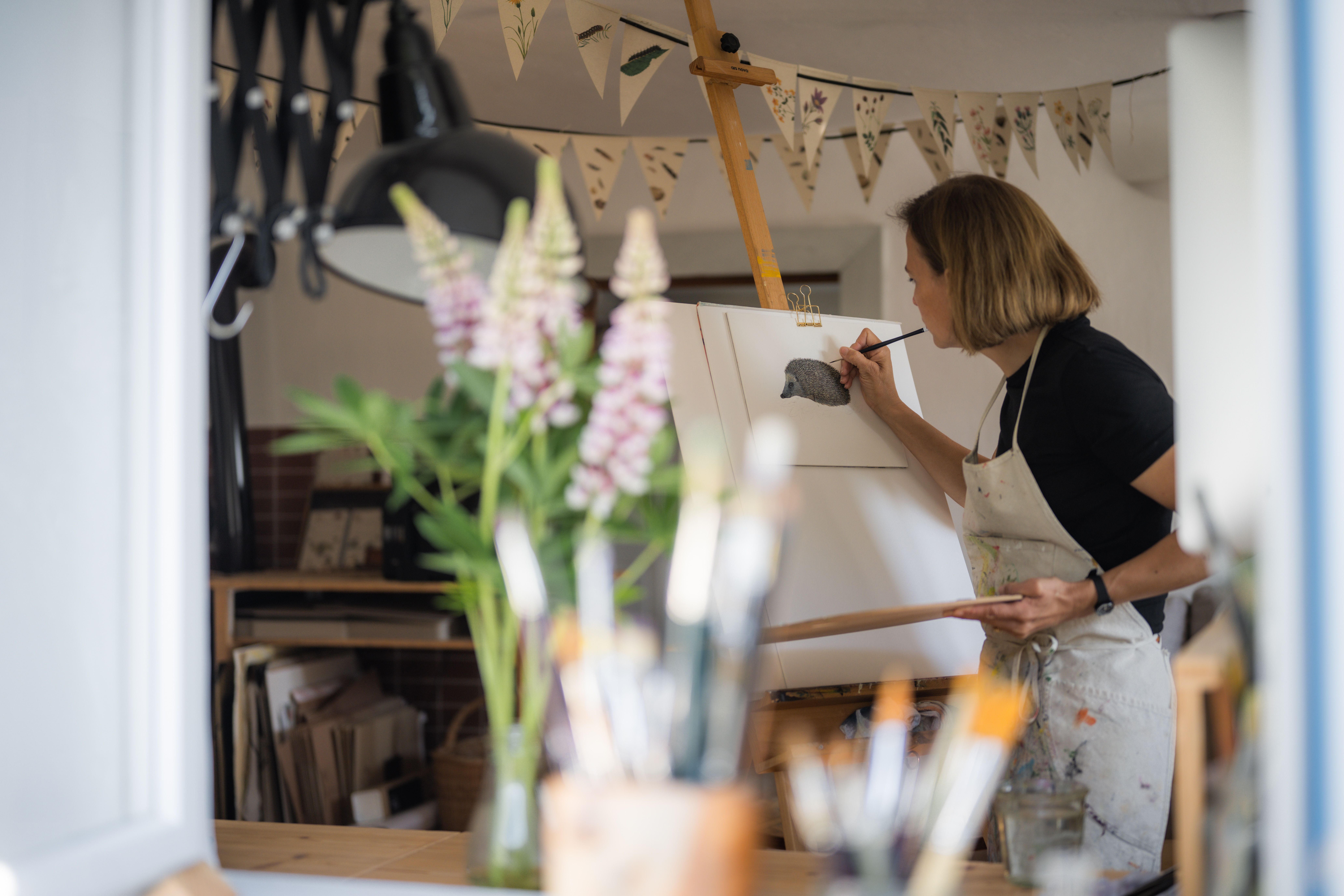 A woman paints a picture of a hedgehog at an easel in a studio. Brushes and flowers can be seen in the foreground.
