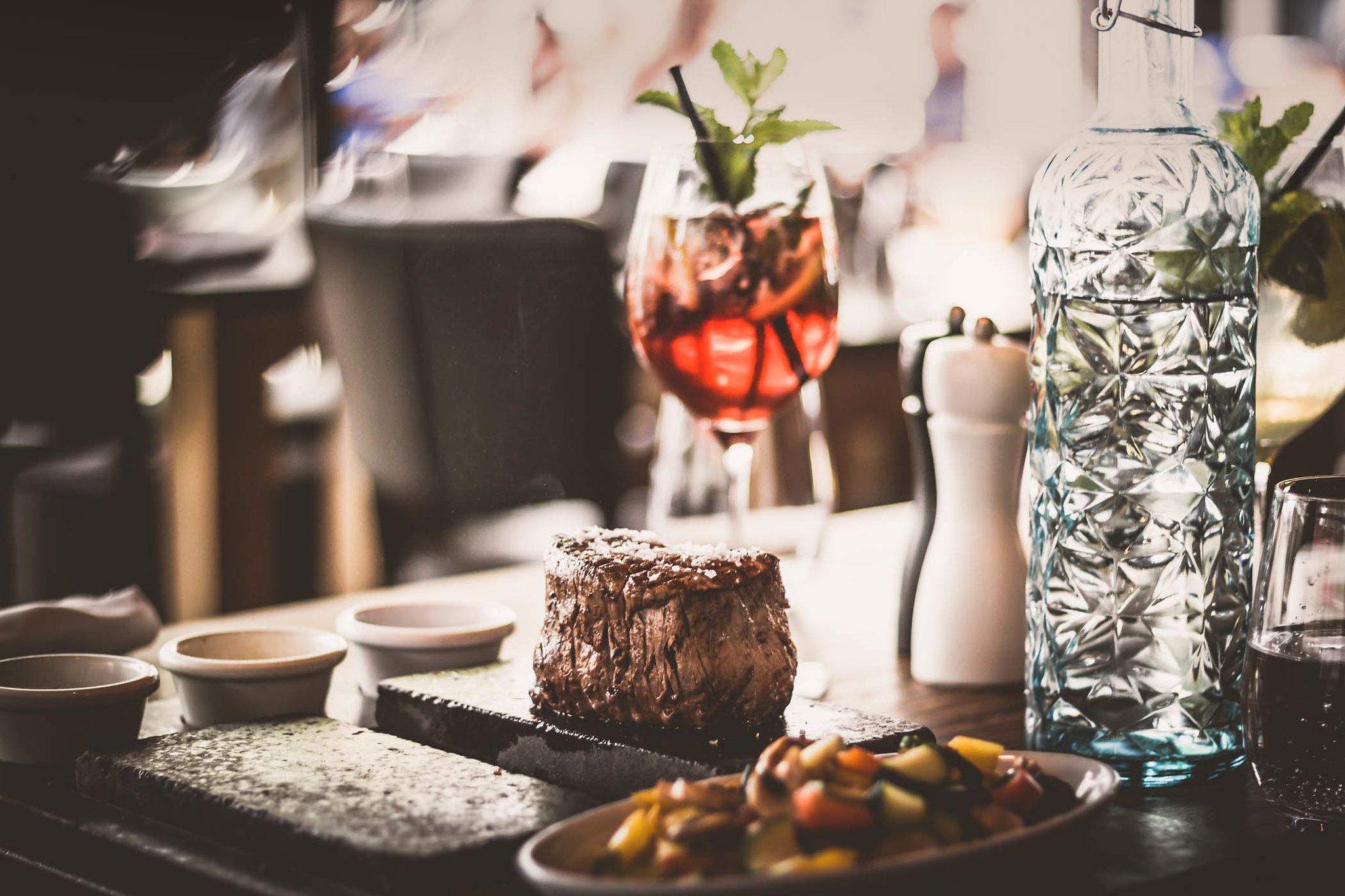 A juicy steak served on a hot stone, accompanied by vegetables, a carafe of water and a cocktail glass.