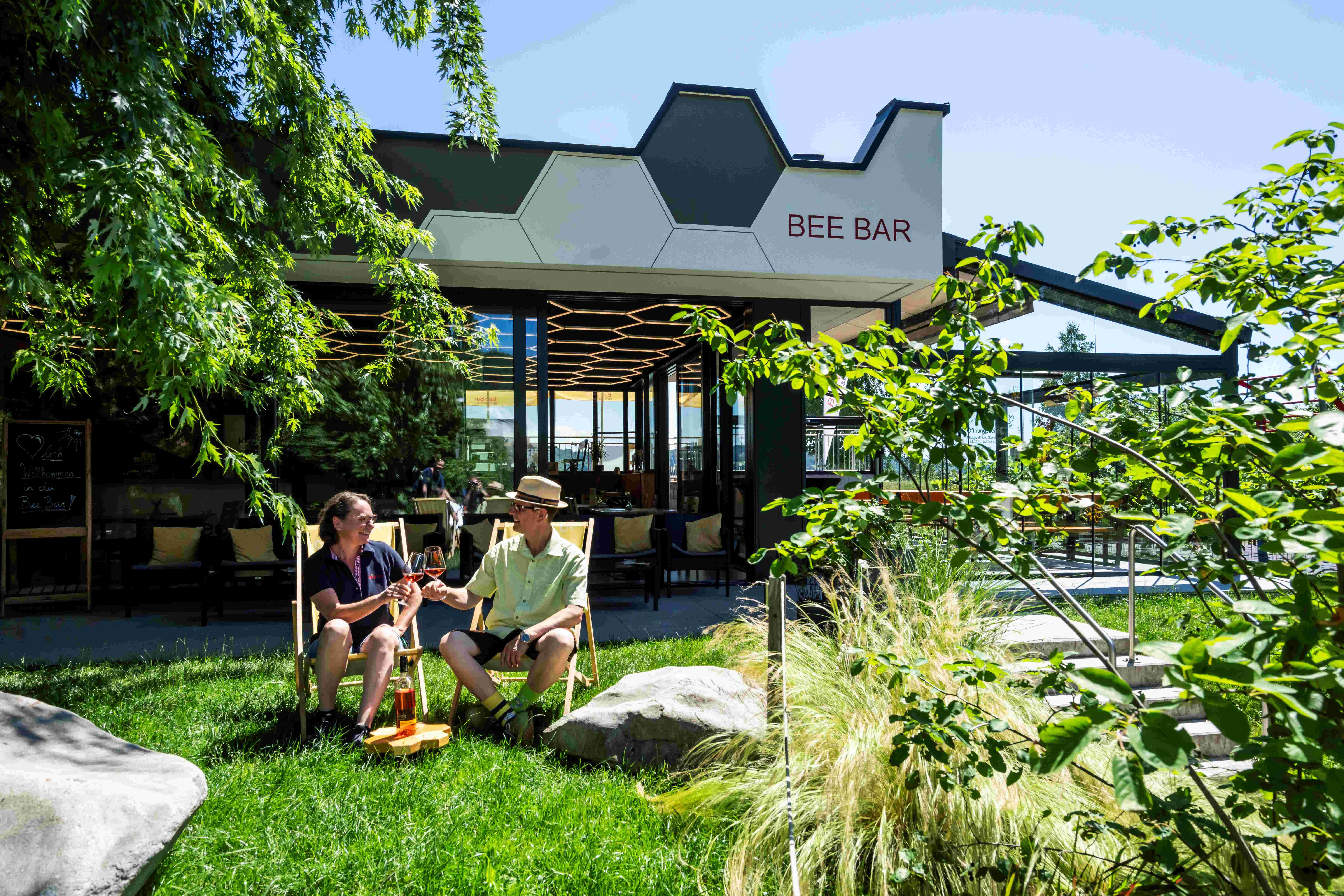 Two people sit in the garden in front of the Bee Bar and toast with drinks.