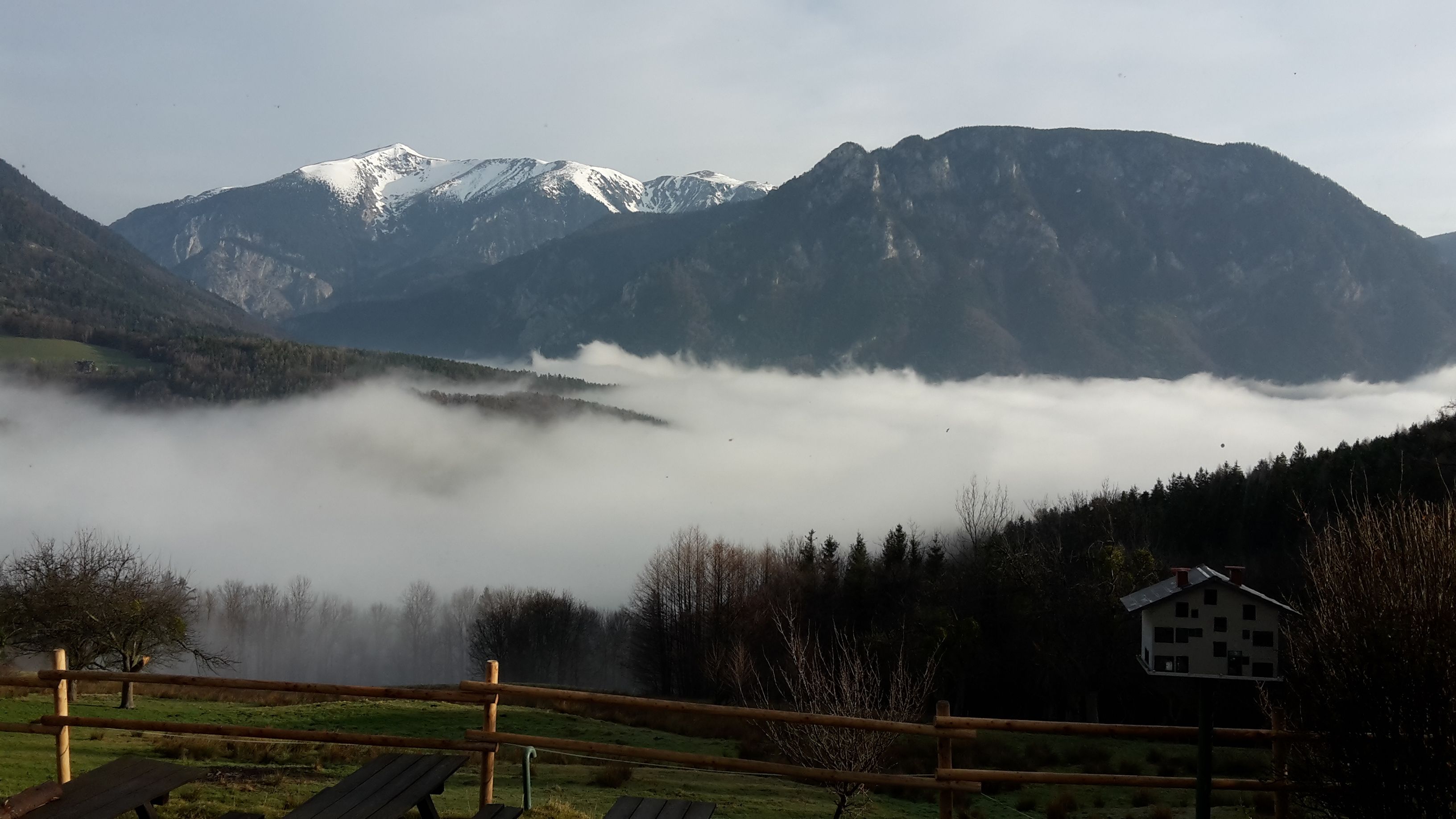 View of snow-covered mountains above a valley with fog and a wooden fence in the foreground.