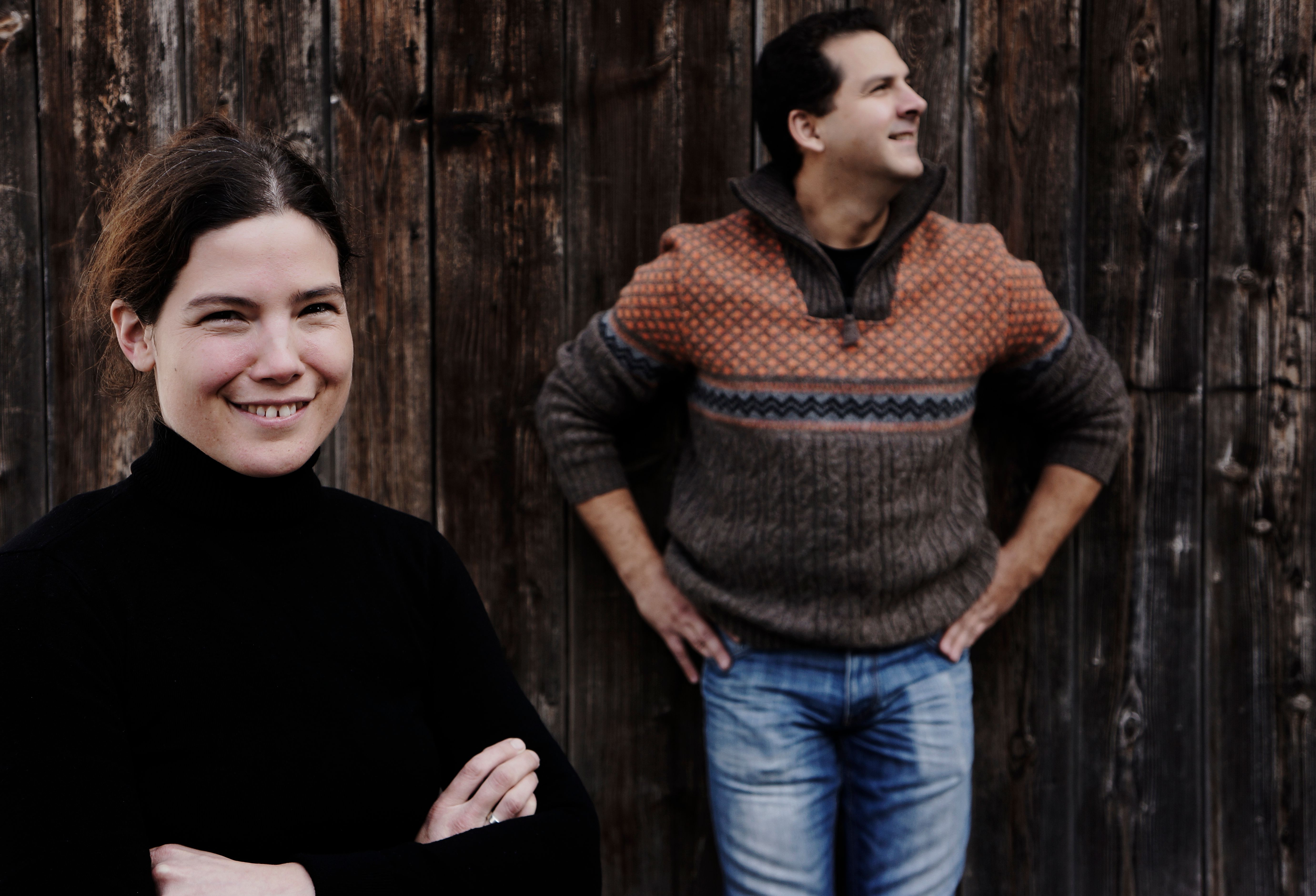 Two people in front of a wooden wall, a woman in the foreground smiling, a man in the background looking to the side.