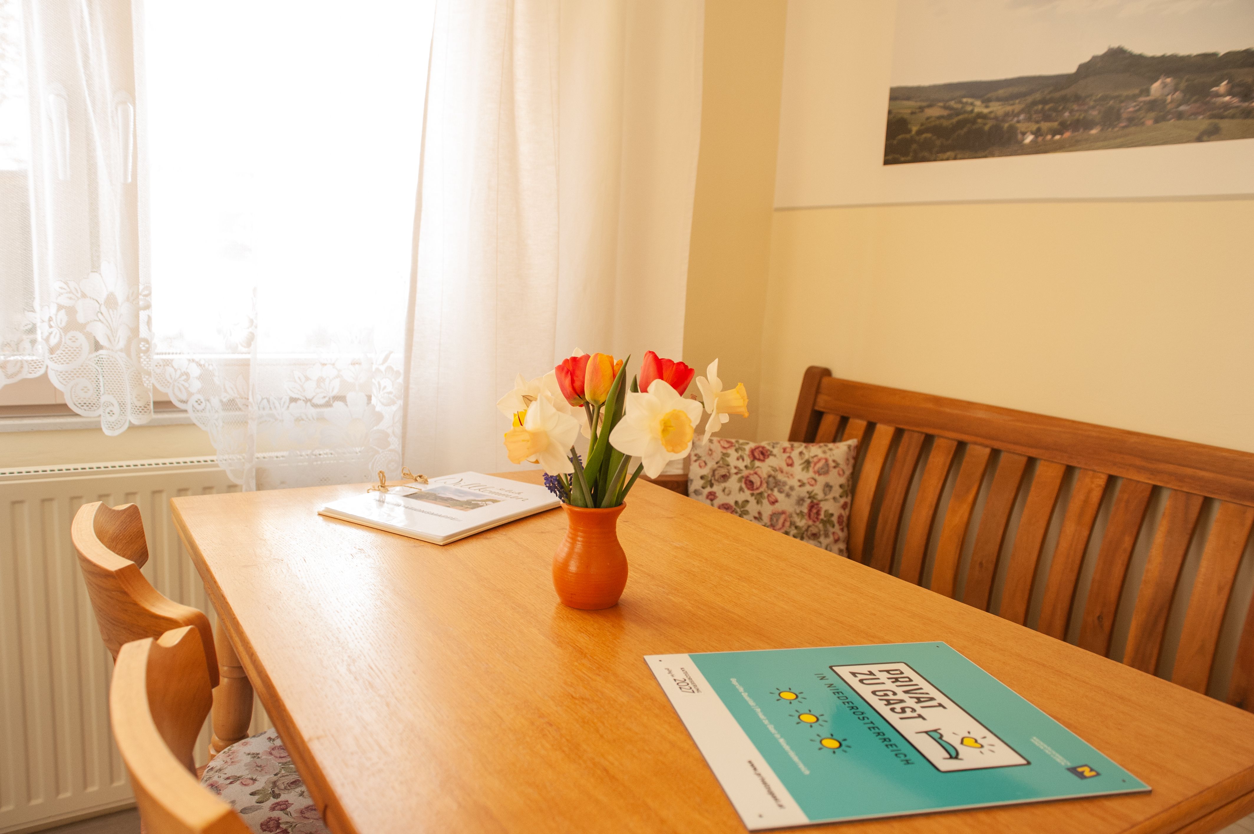 A cozy dining area with a wooden table, a bouquet of flowers in a vase and brochures. Light-colored curtains and a picture on the wall.