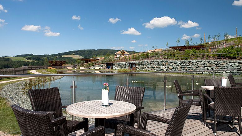 A wooden terrace with round tables and chairs in front of a pond.