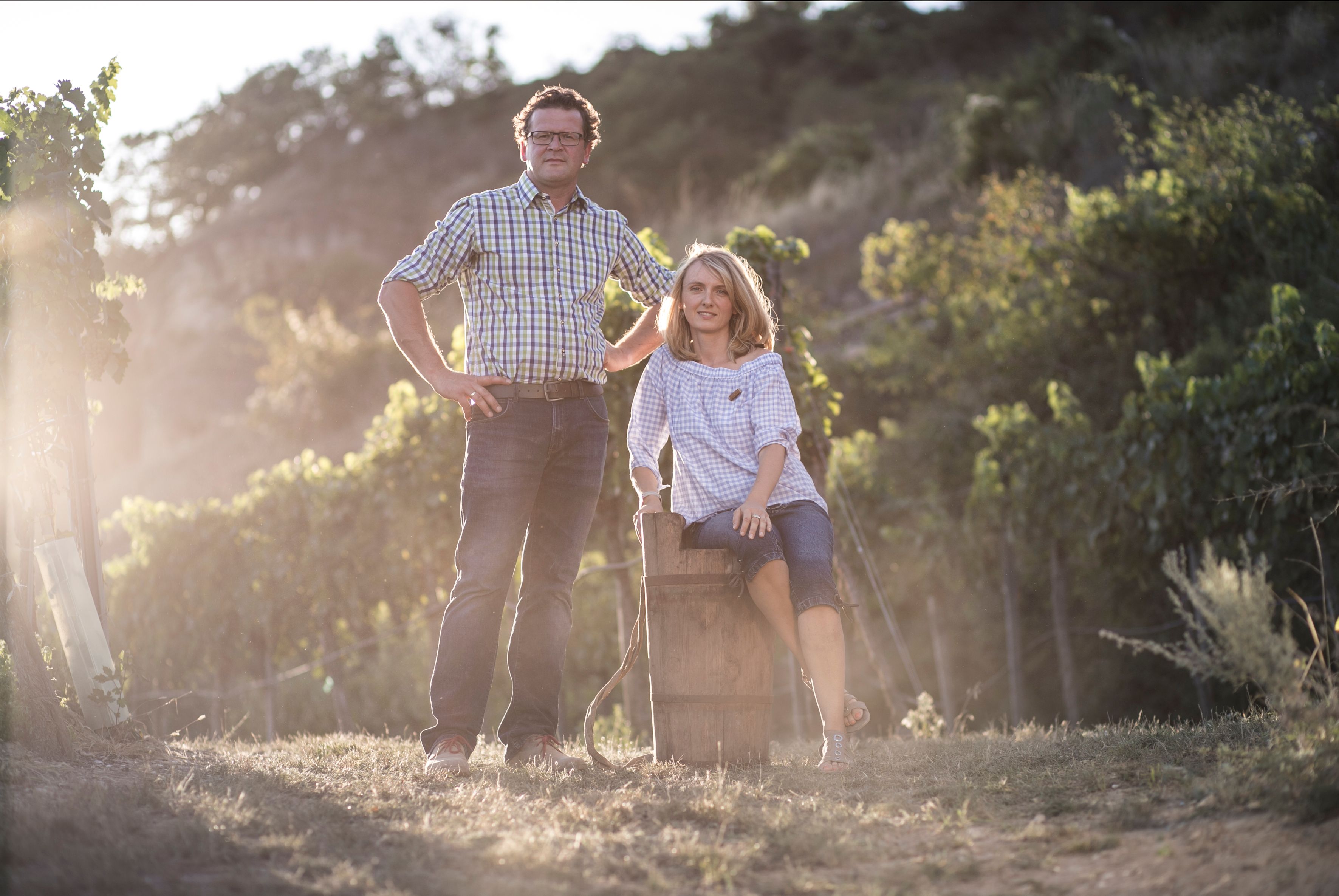 A man and a woman are standing in a vineyard, the man standing and the woman sitting on a barrel. The sun is shining through the leaves.
