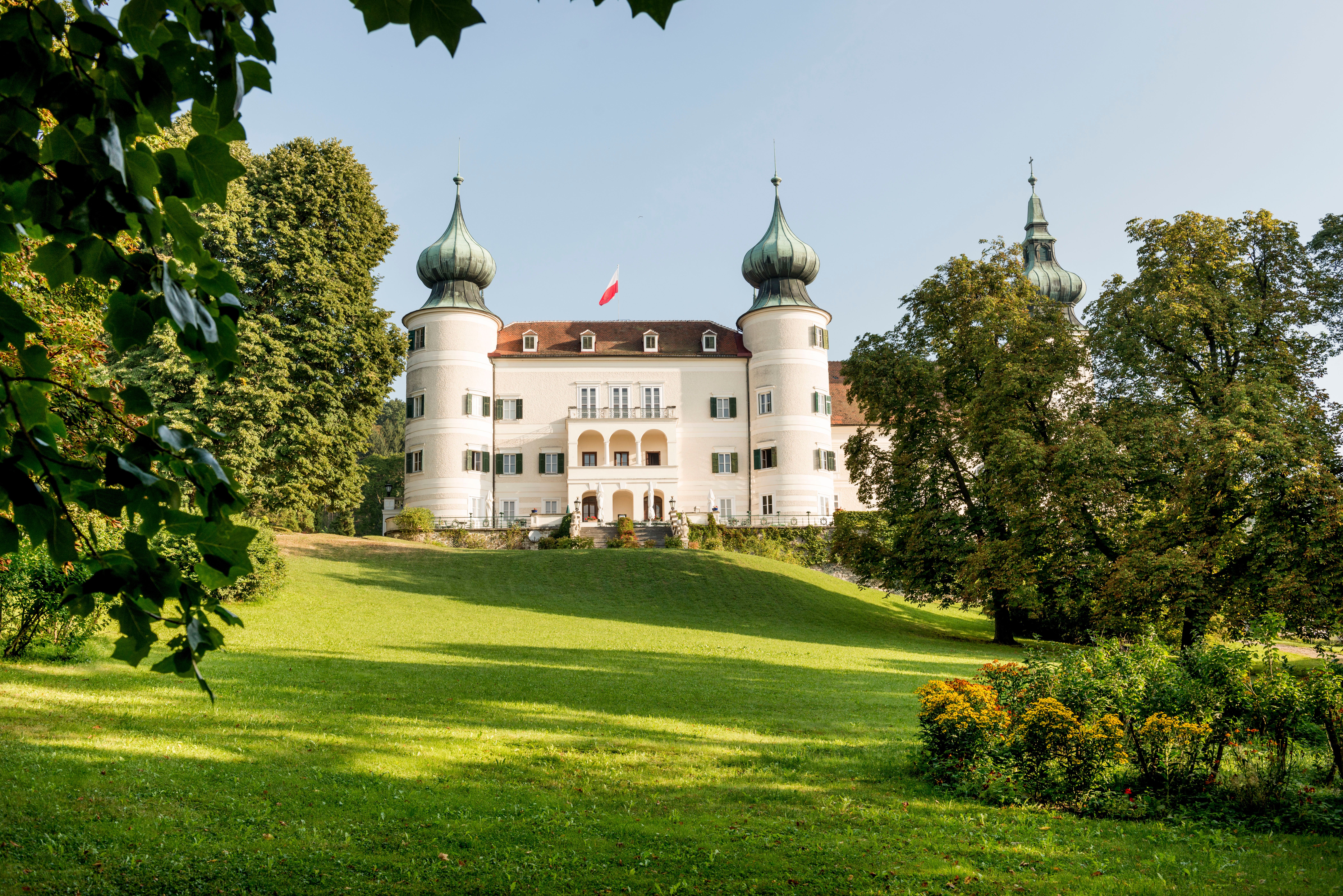 A white castle with several onion domes, surrounded by green countryside and trees, under a blue sky.