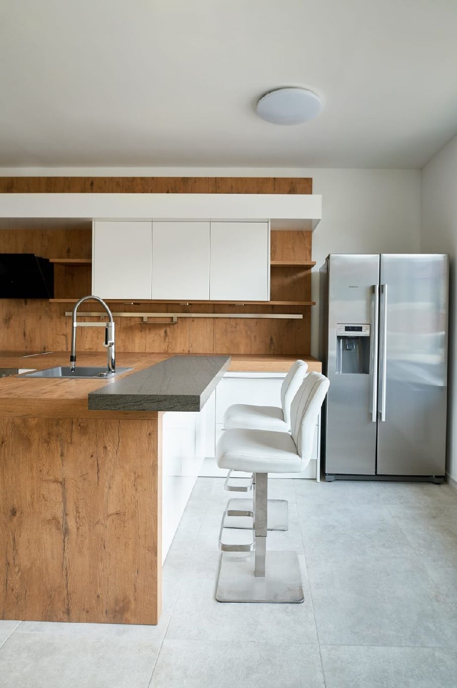Modern kitchen with wood and white tones, bar stools and stainless steel fridge.