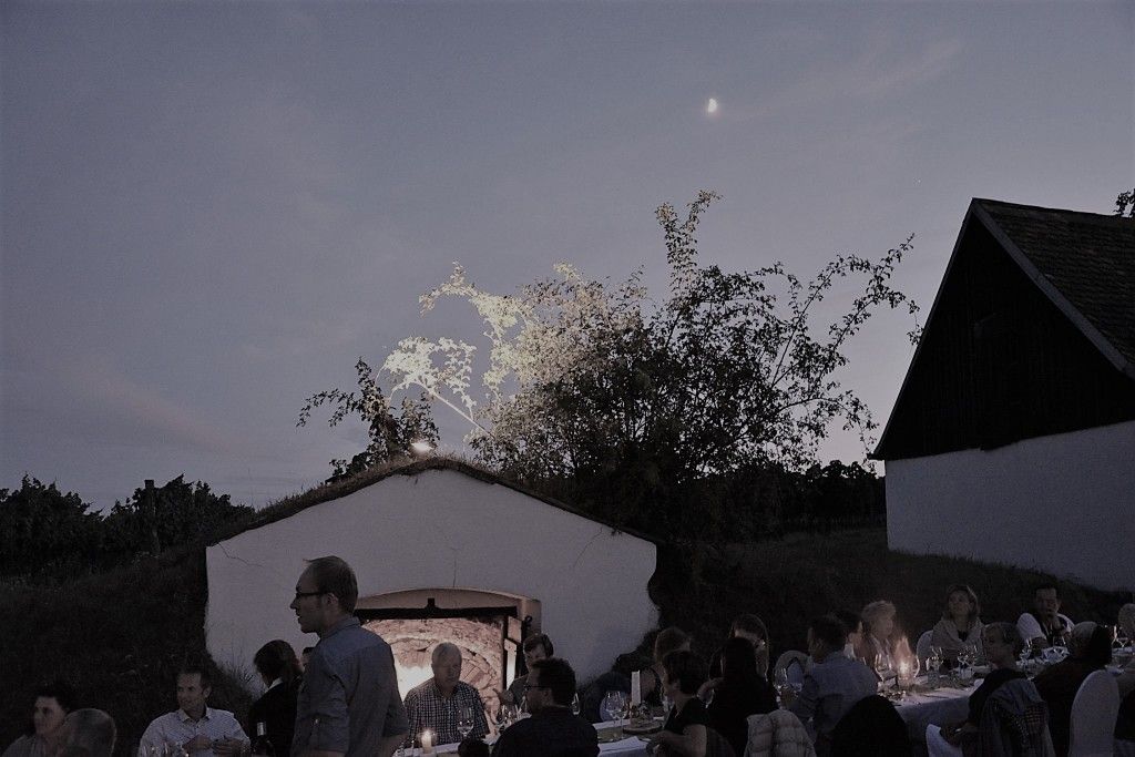 People sit at a long table outside at dusk, surrounded by buildings and trees.