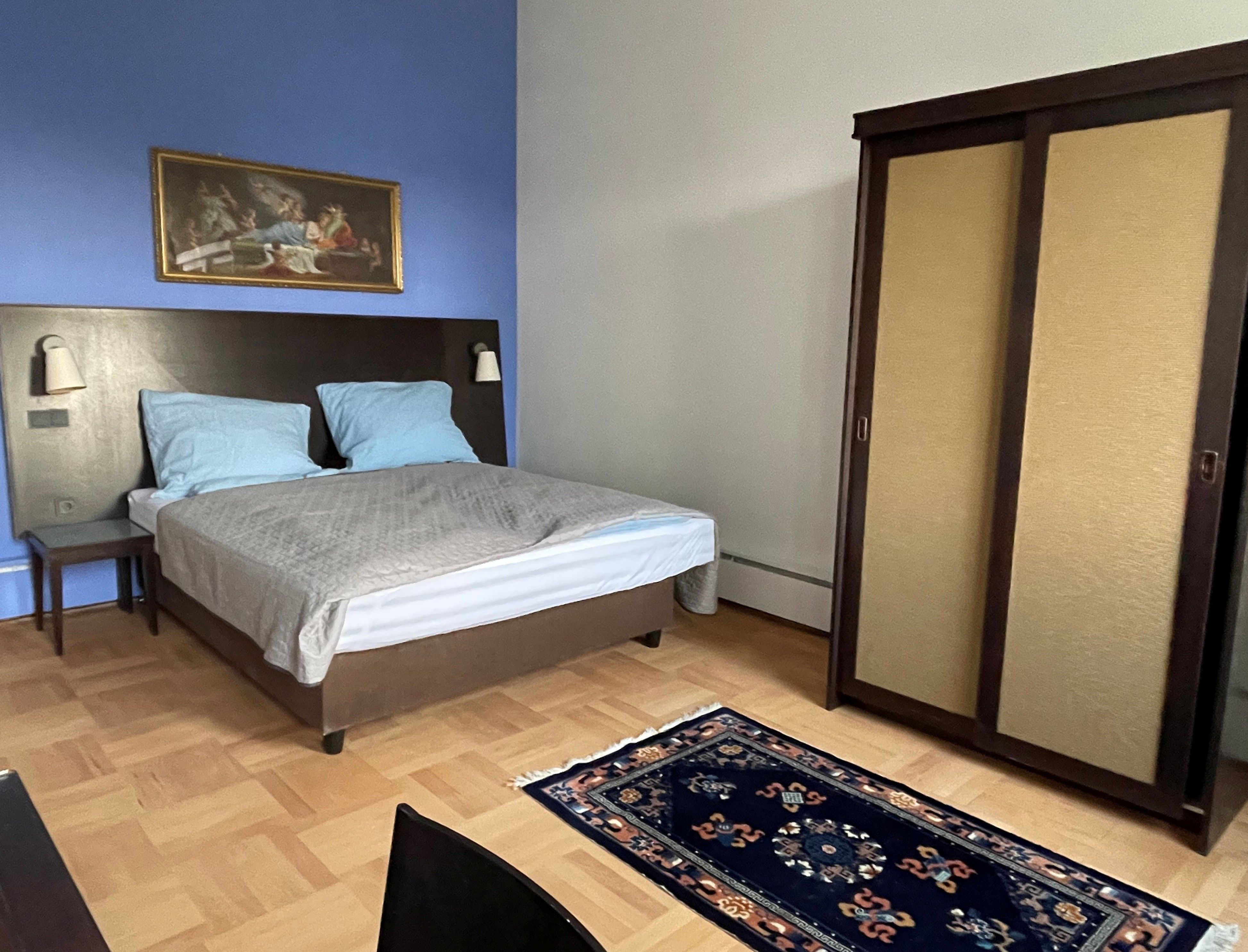 A bedroom with a blue wall, a double bed, a closet, and a rug on a wooden floor.
