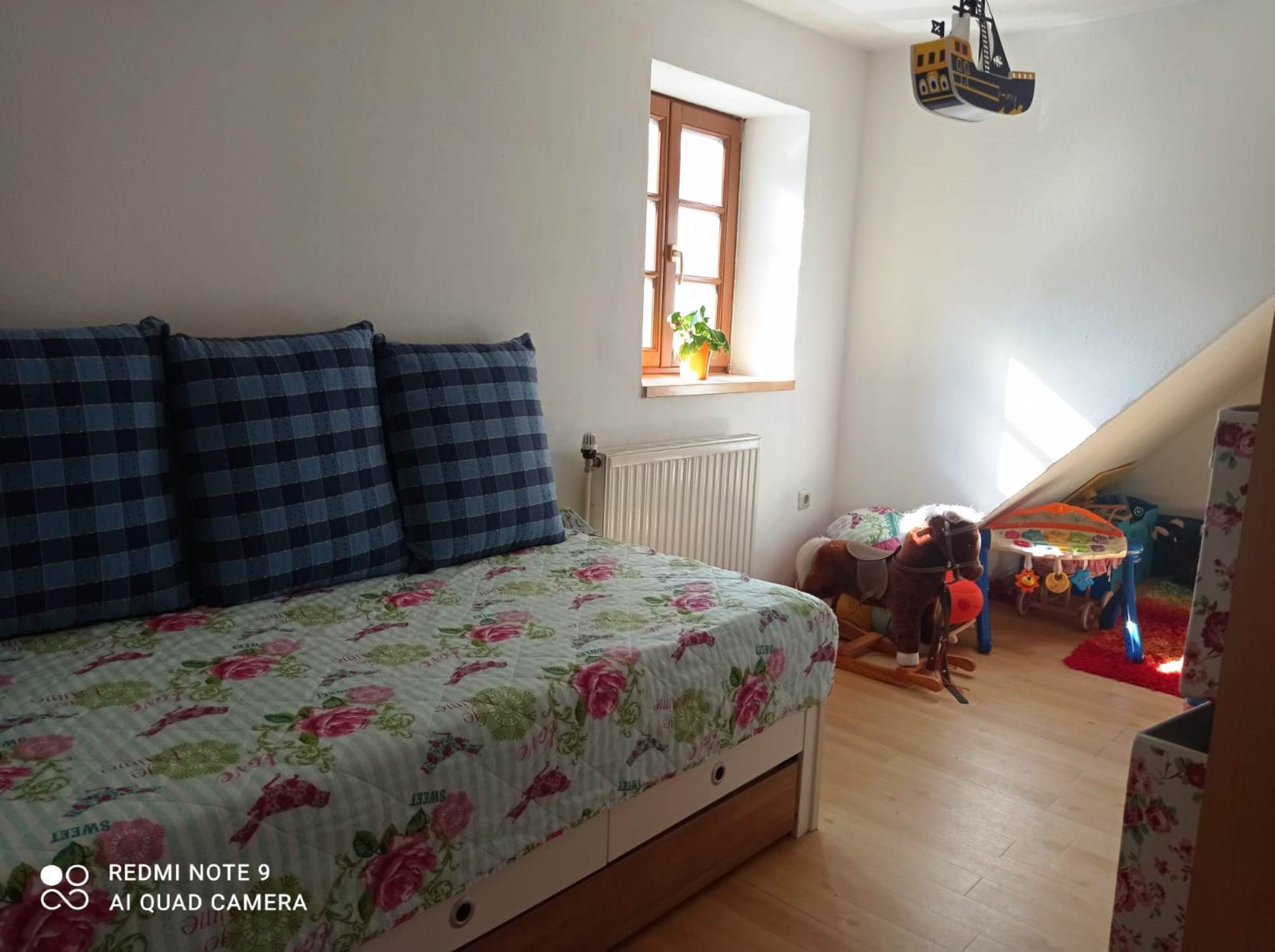 Children's room with pull-out bed, plaid cushions, toy horse and play table. Bright window with plant.