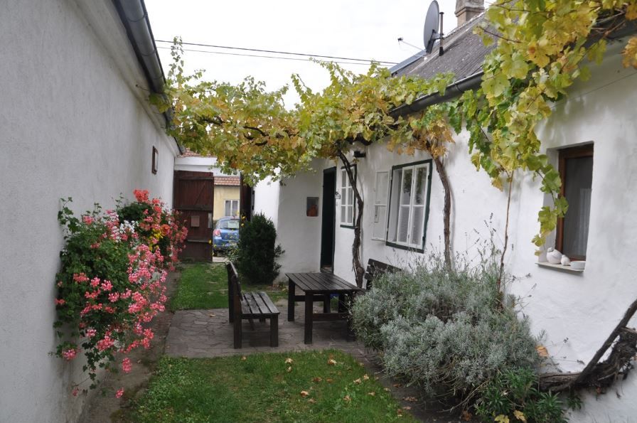 A cozy courtyard with a wooden table and benches, surrounded by white walls, flowers and vines.