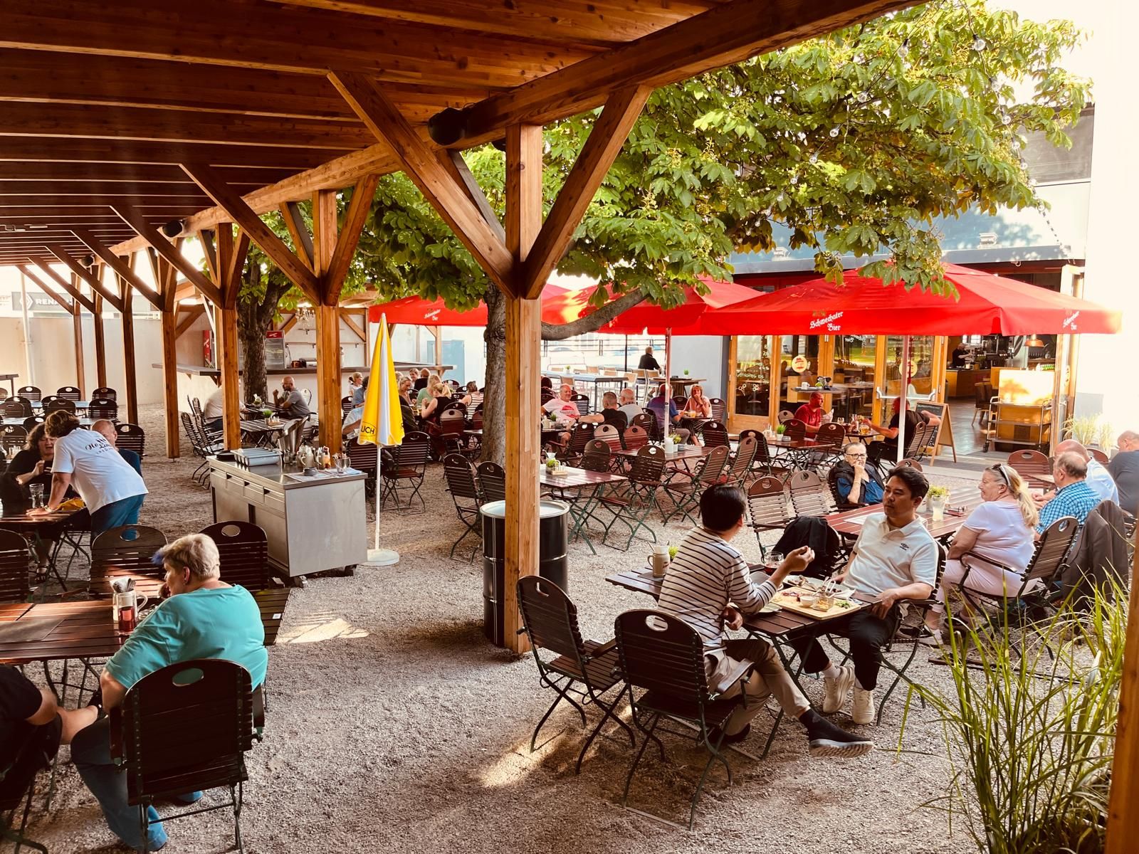 A beer garden with a wooden roof, red parasols and lots of people at tables.