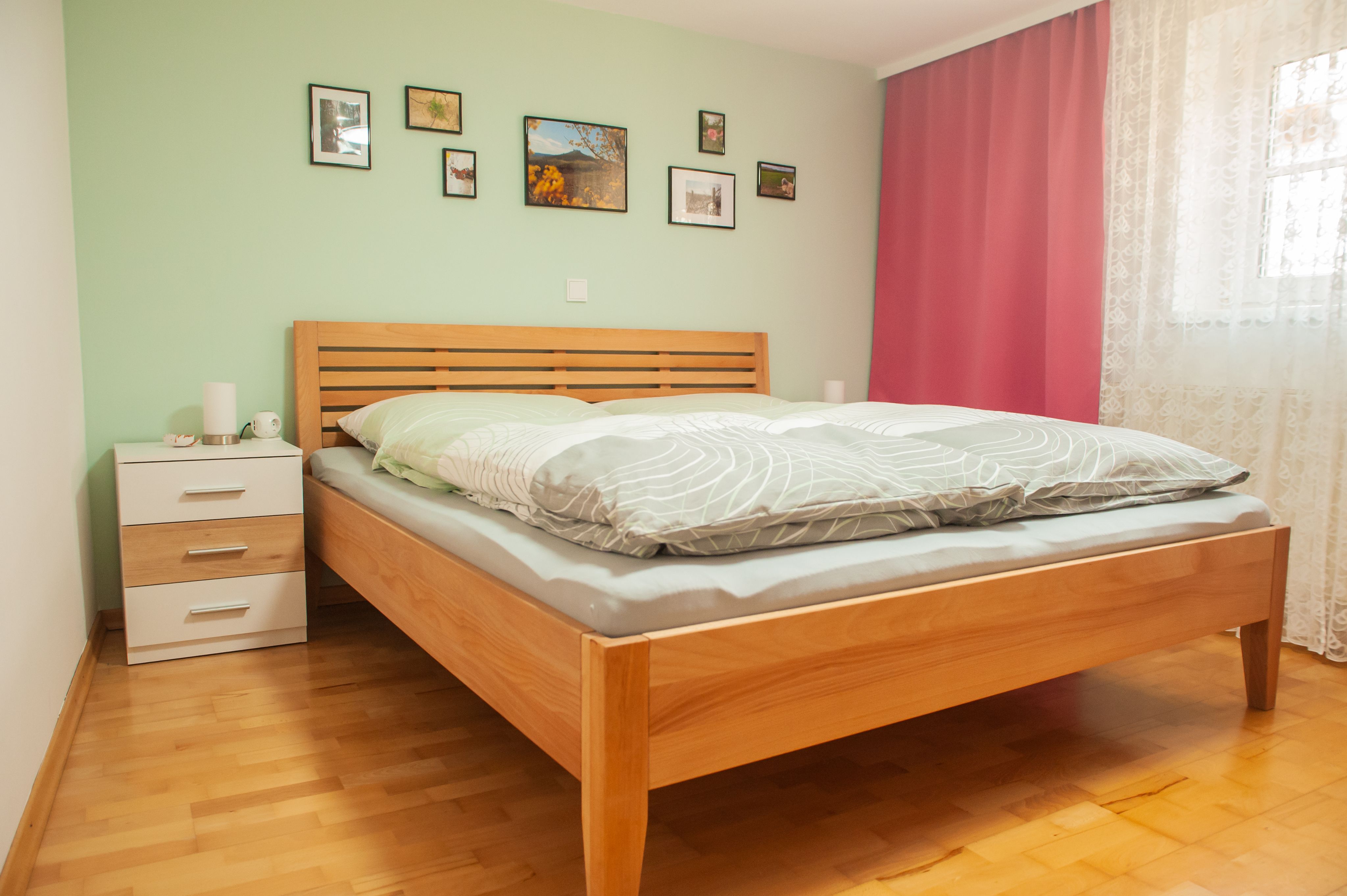 A bedroom with a wooden bed, green wall, pictures, bedside table and pink curtains.
