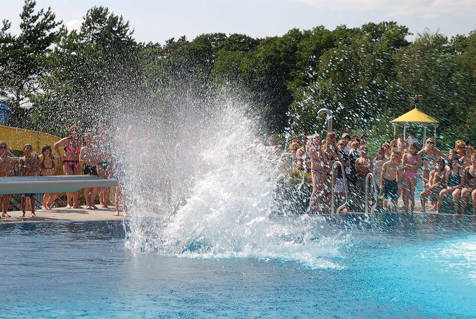 People in an outdoor pool watch a large splash of water in the pool.