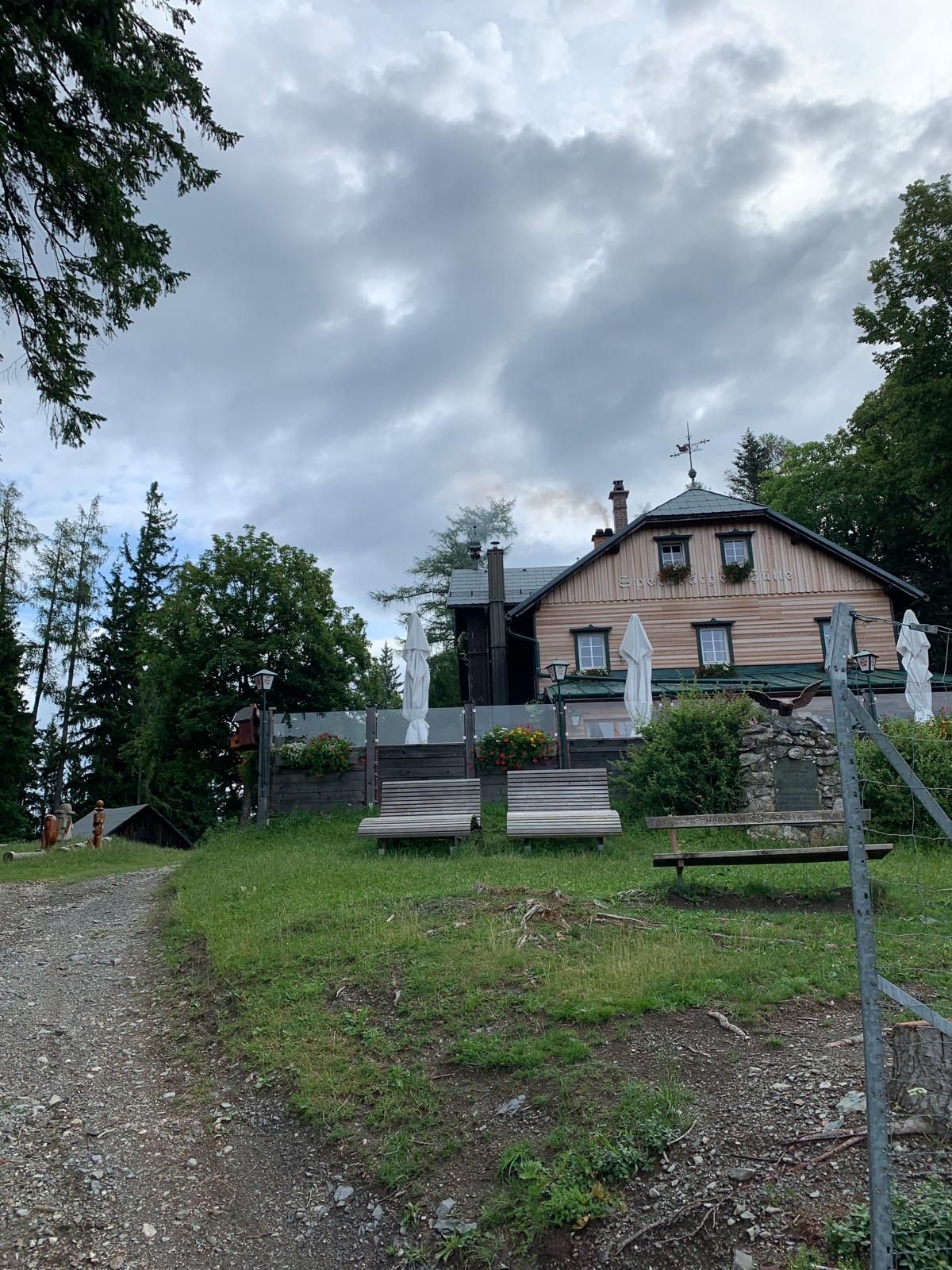 A mountain hut with a terrace and deckchairs on a meadow, surrounded by trees and a cloudy sky.