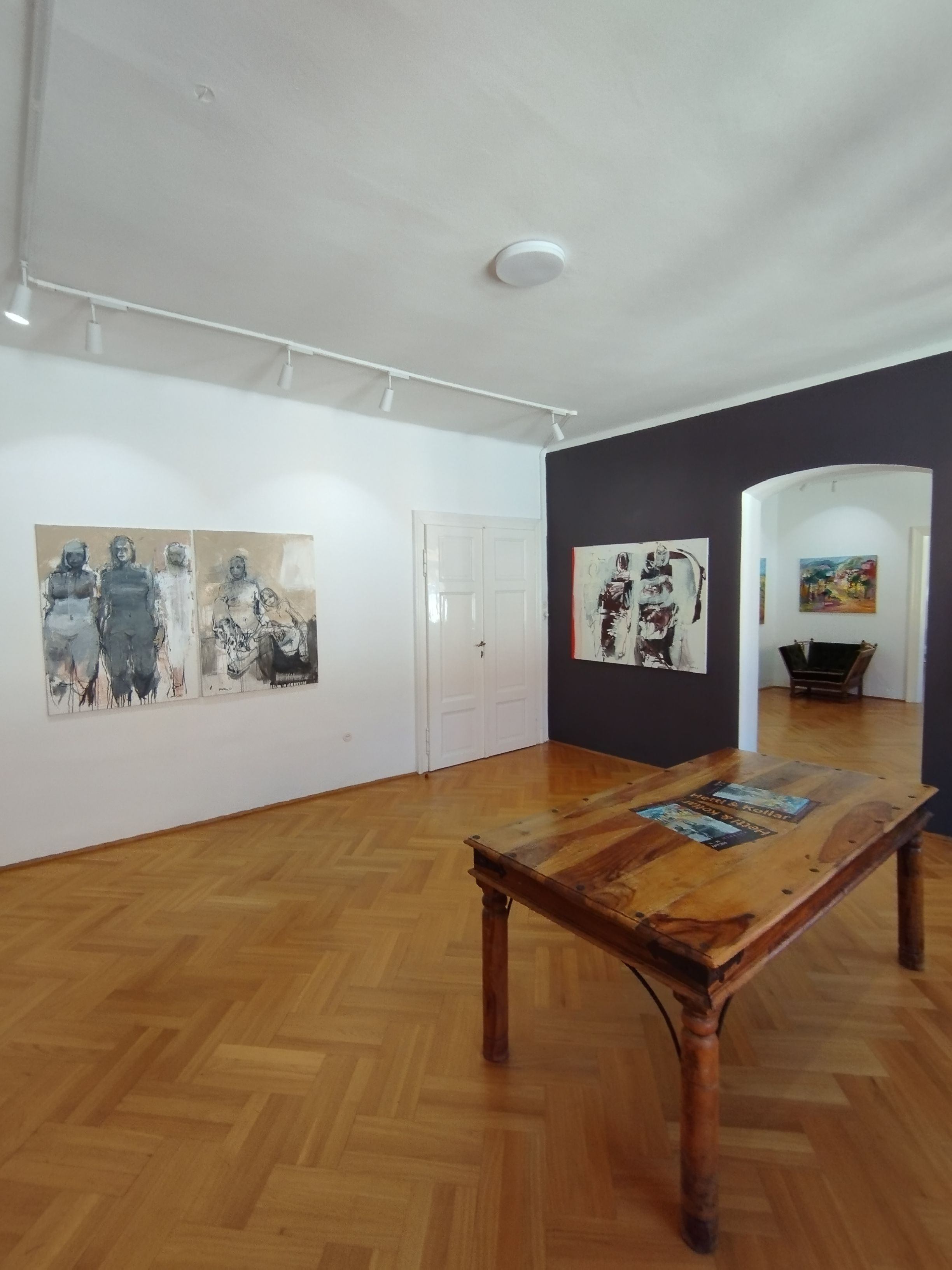 Interior view of an art gallery with a wooden floor, paintings on the walls and a wooden table in the middle.