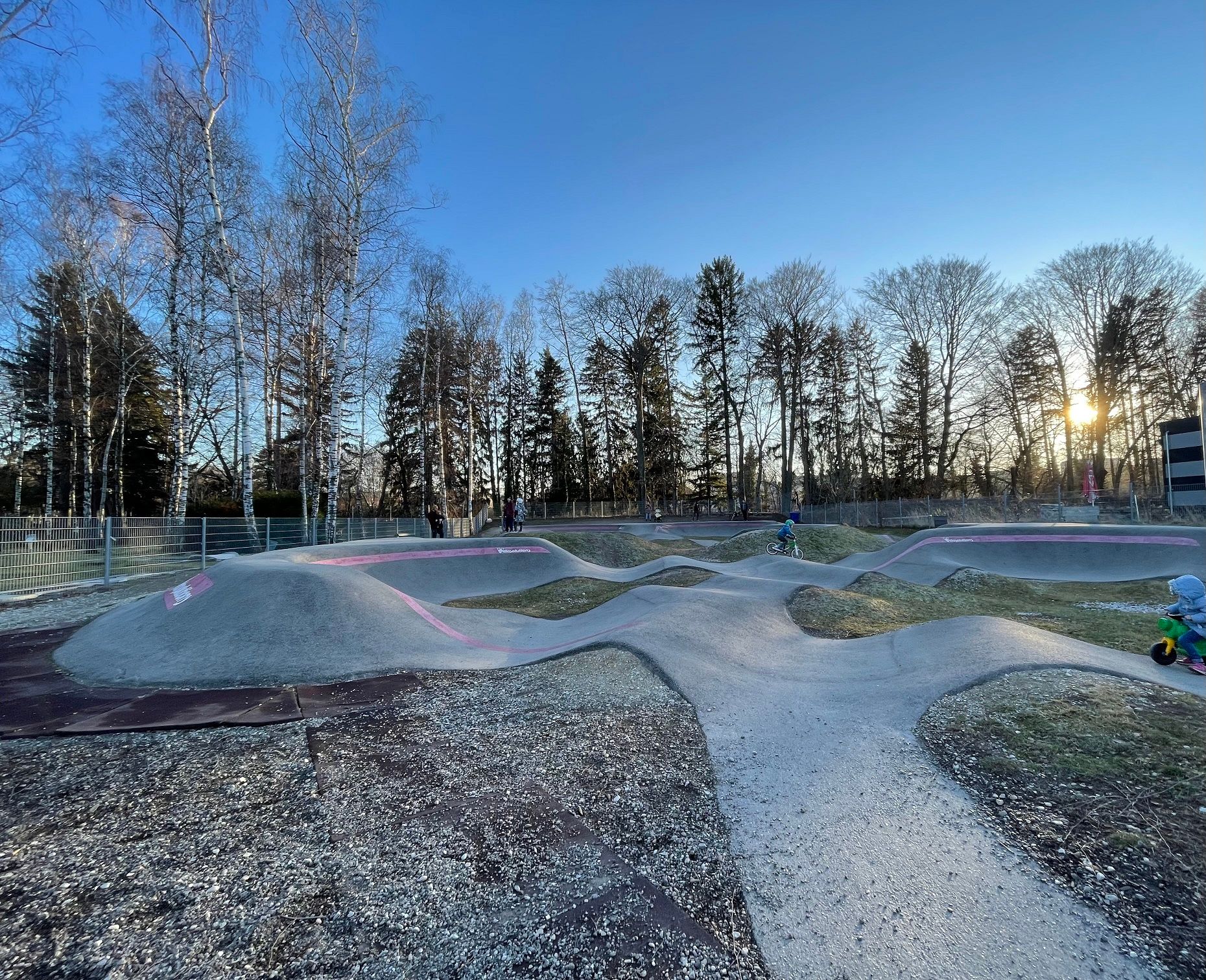 Pump track in Neunkirchen with children on bikes and balance bikes, surrounded by trees at sunset.