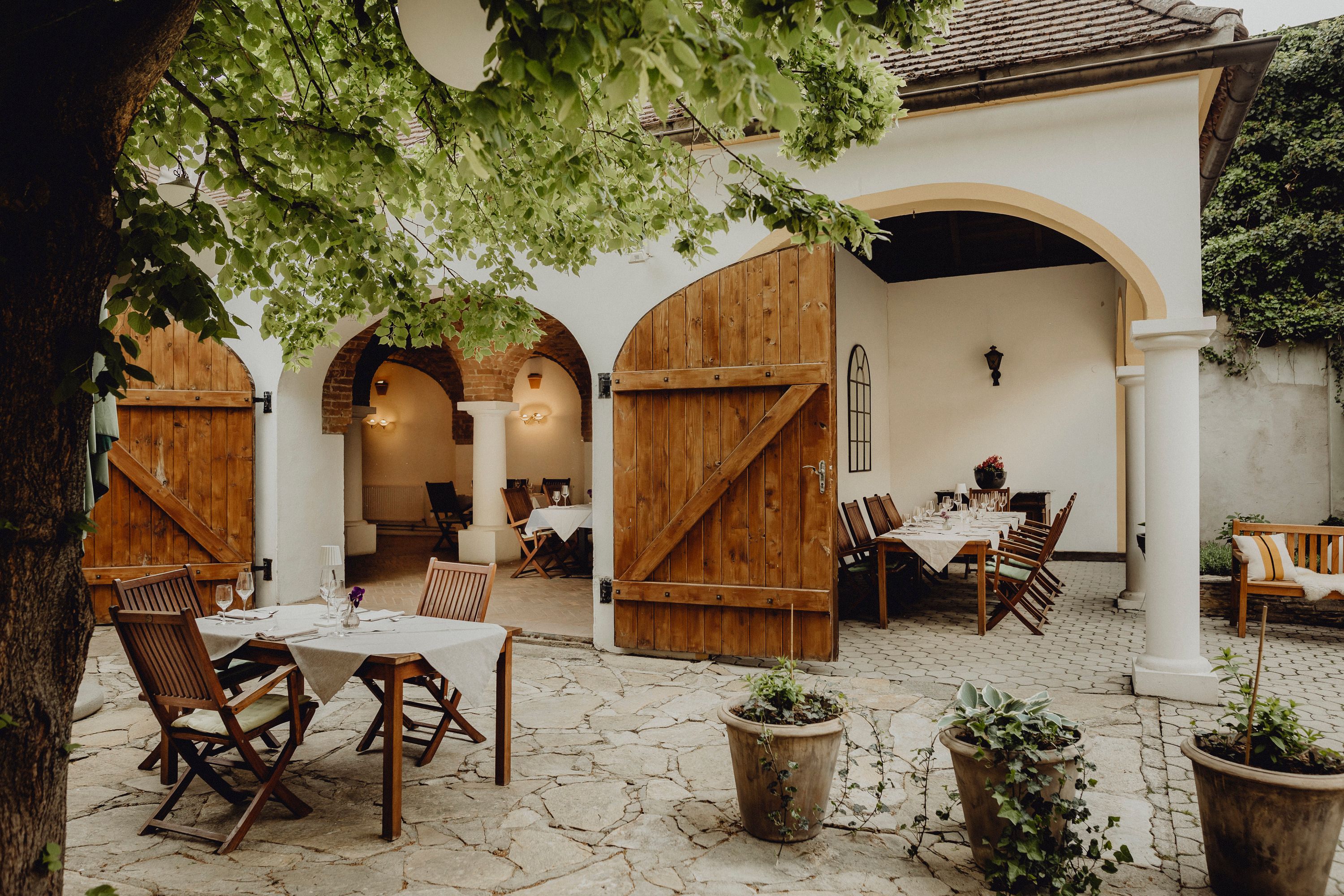 Cozy guest garden with wooden tables and chairs under a tree, surrounded by white walls and open wooden doors.