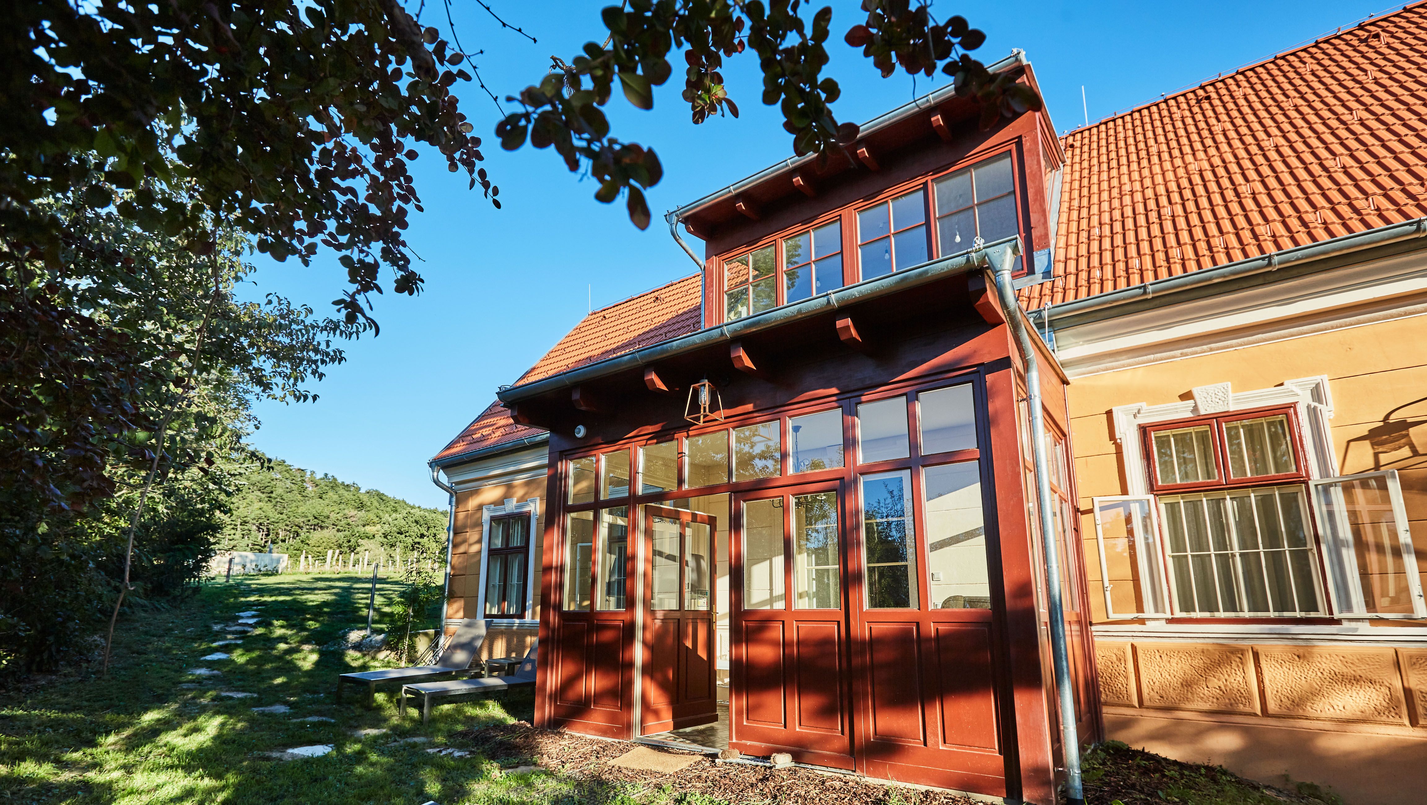A yellow villa with red roof tiles and a glazed entrance, surrounded by trees and a green garden.