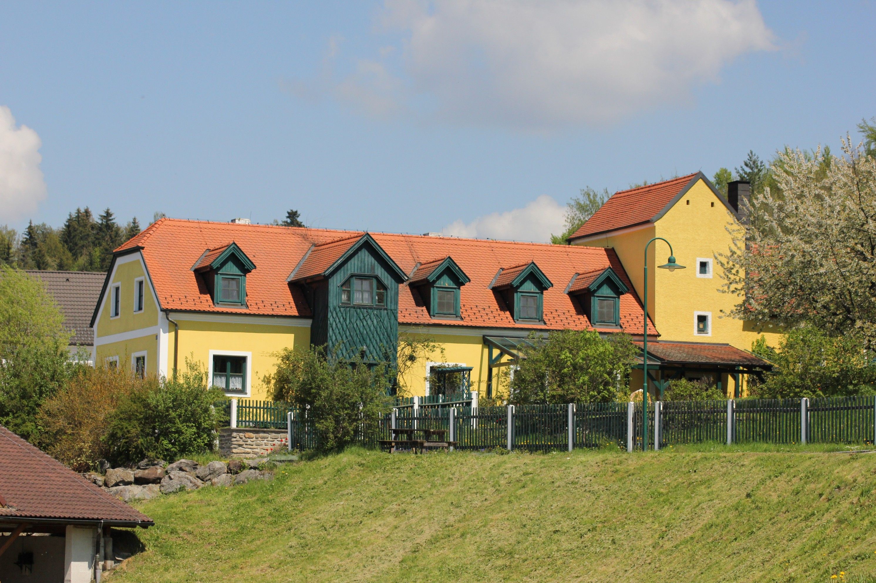 Yellow house with a red roof and green shutters, surrounded by trees and a fence.