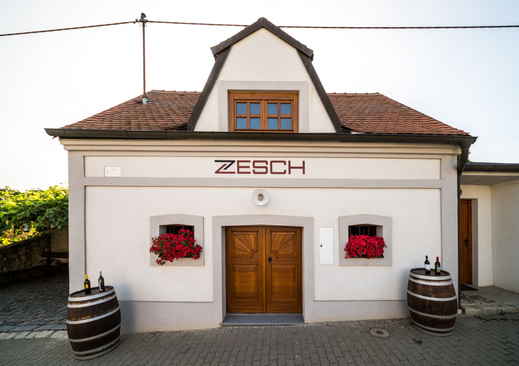 Small building with the inscription 'Zesch', two wine barrels and red flowers.
