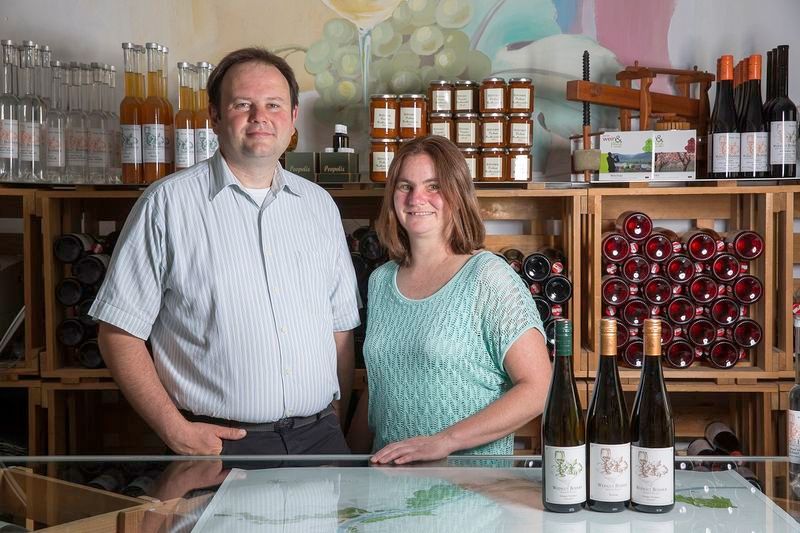 Two people stand in front of wine bottles and shelves of products in a winery.