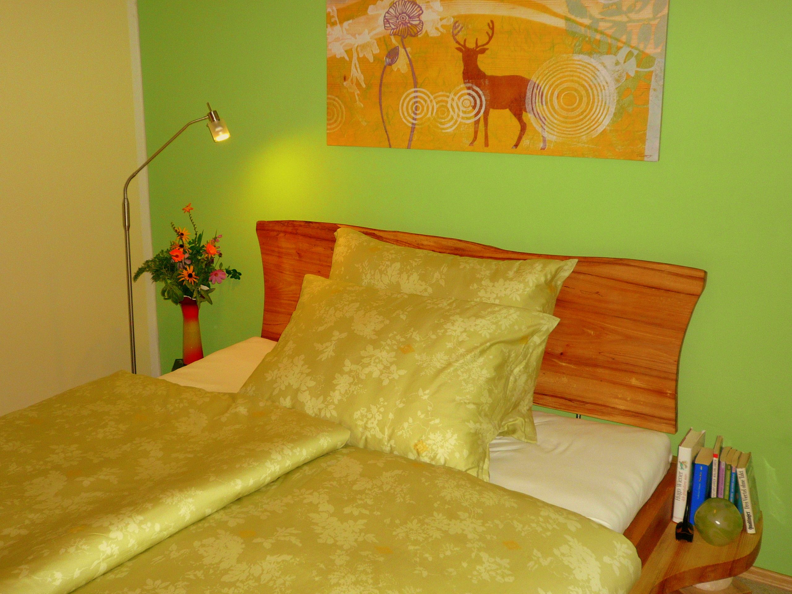Cozy bedroom with green bedding, wooden headboard, green wall and deer picture. There is a lamp and flowers on the bedside table.