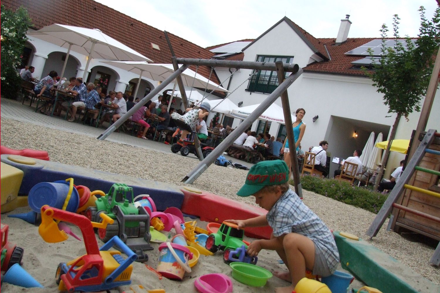 A child plays in a sandpit with colorful toys. In the background, people sit at tables under parasols.