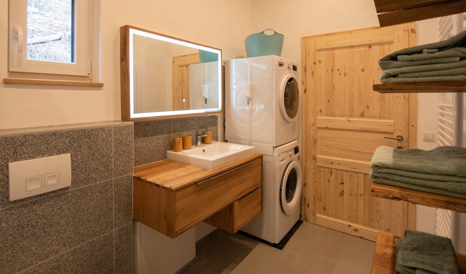 Modern bathroom with wooden washbasin, illuminated mirror, washing machine and dryer. Towels on wooden shelves.
