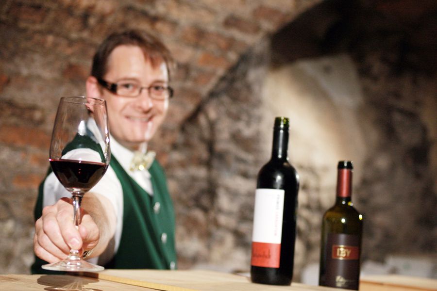 A man in traditional dress offers a glass of red wine, with two bottles of wine in the background.
