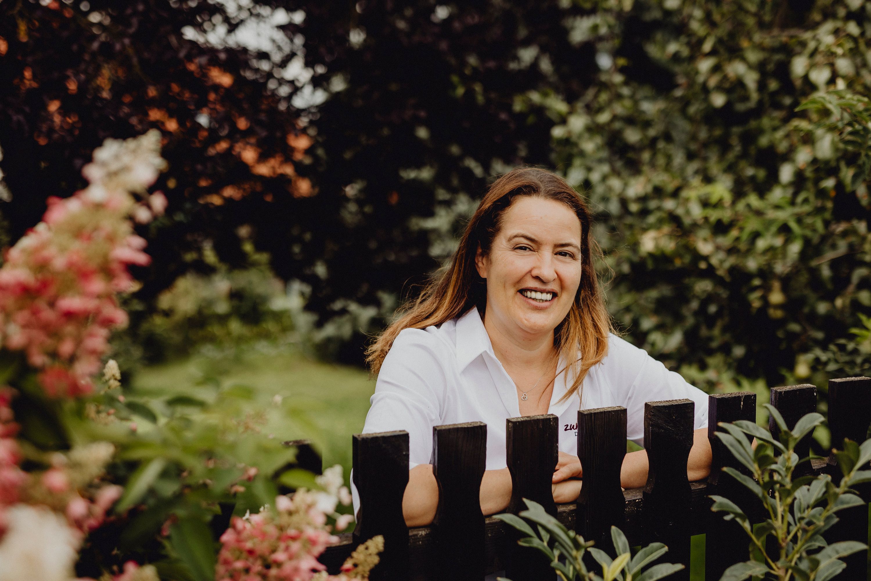 Woman in white shirt smiling behind a wooden fence in the garden.