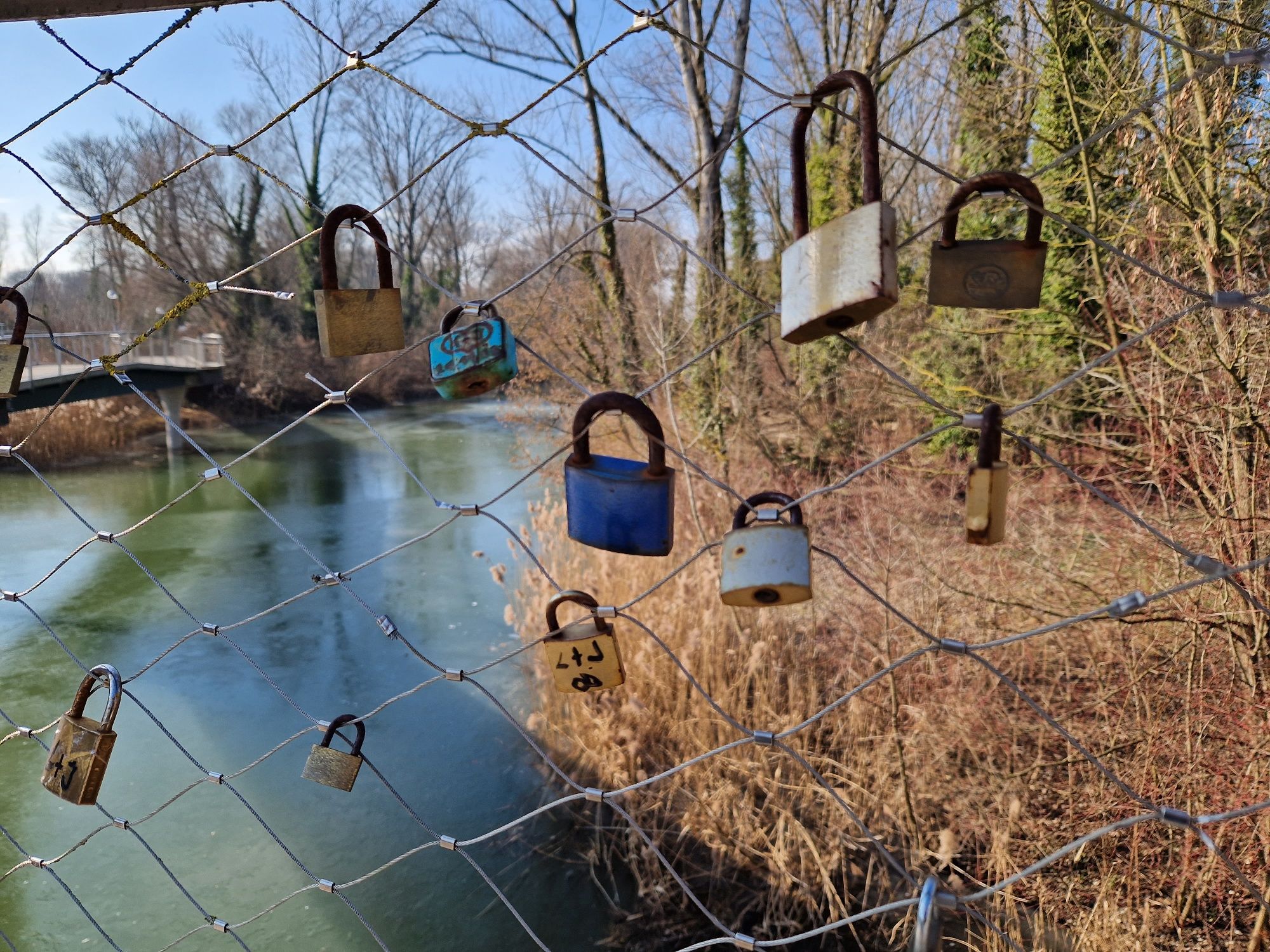 Several padlocks on a wire mesh fence over a river.