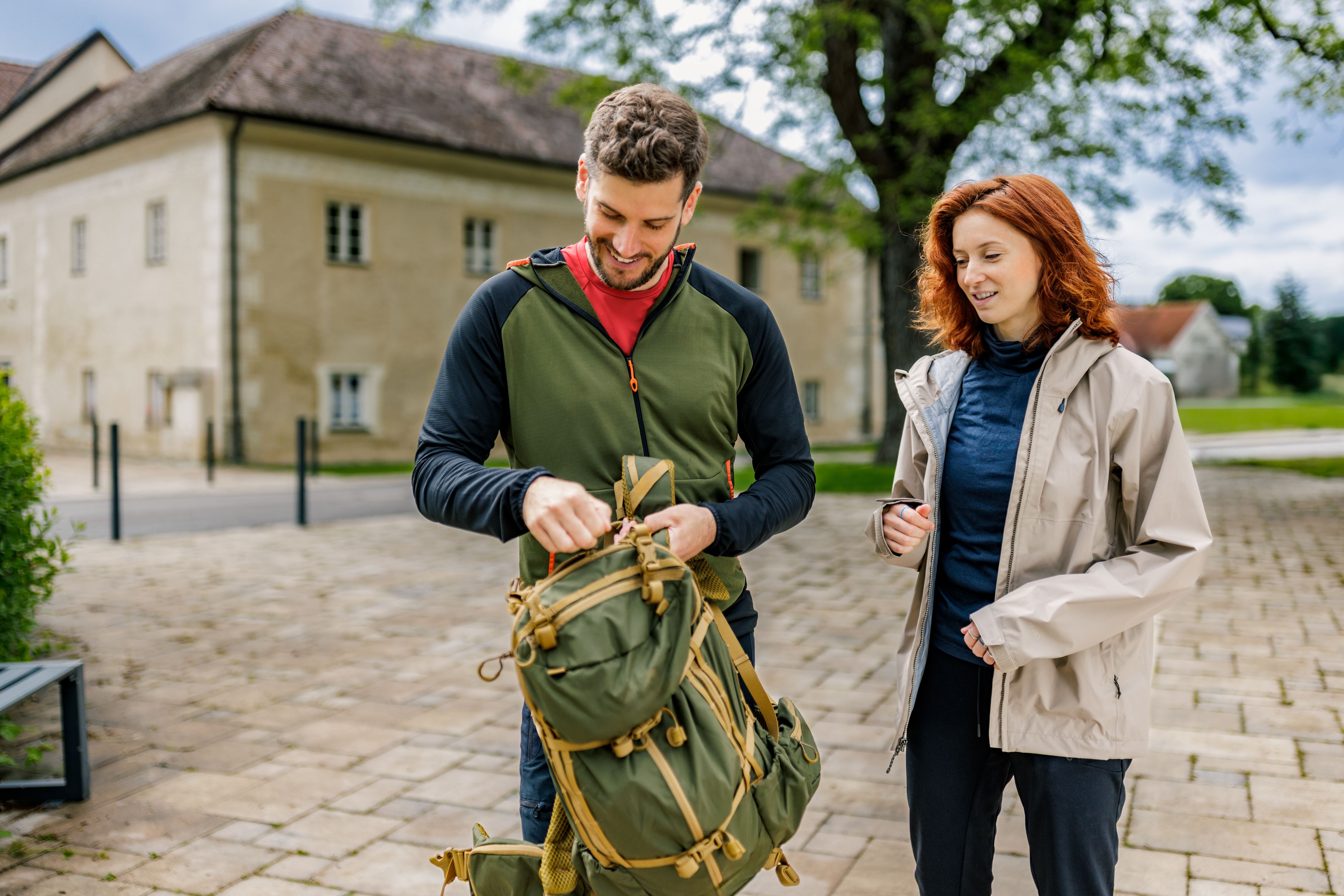 A man and a woman are standing outside Katzelsdorf Castle. The man is packing a green rucksack while the woman looks on. Both are wearing outdoor clothing.