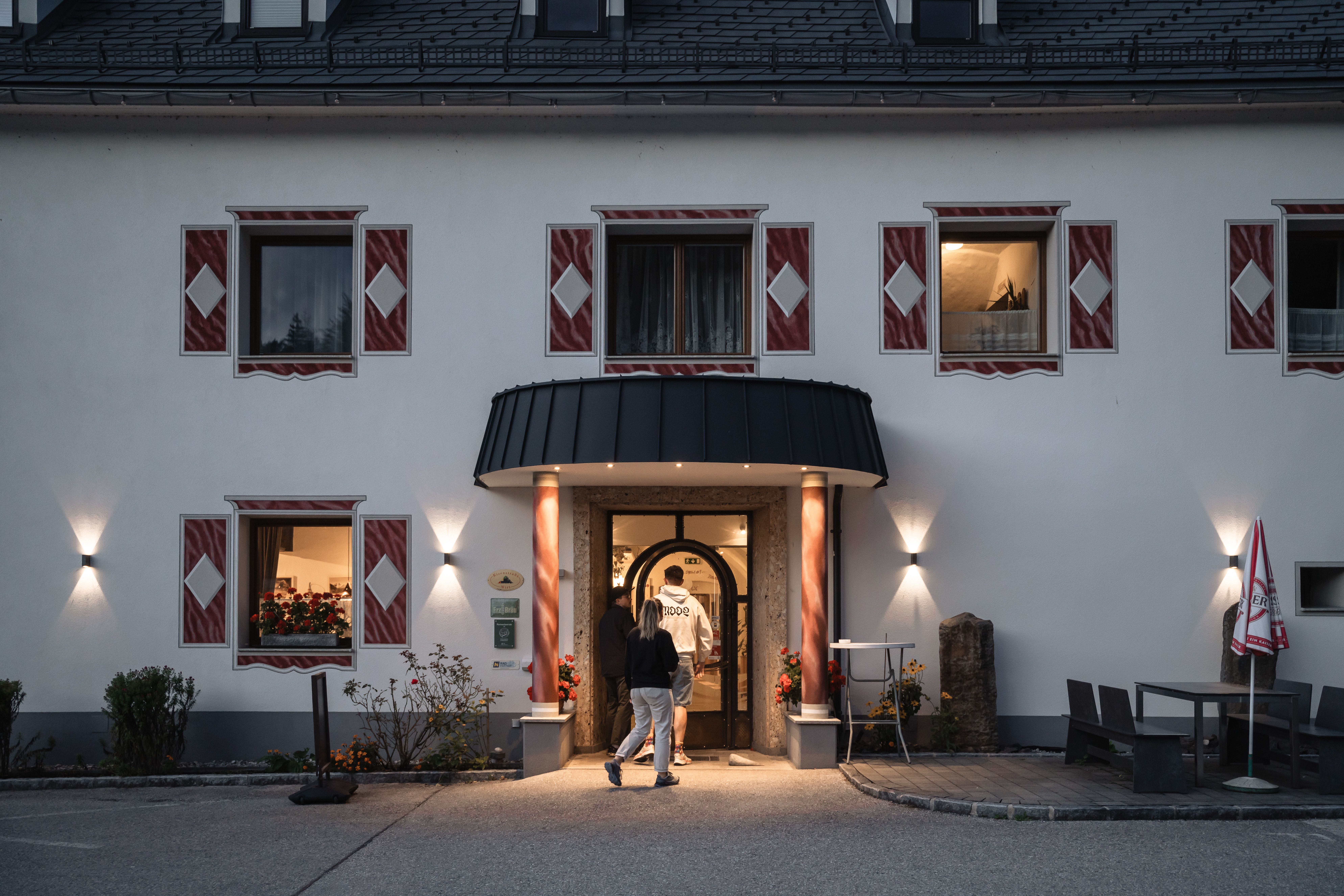 Der Eingangsbereich des Landhotels und Restaurants Zellerhof in Lunz am See bei Abenddämmerung. Drei Personen betreten das Gebäude durch eine einladend beleuchtete Tür, eingerahmt von dekorativen Fensterläden. Die Beleuchtung und das äußere Erscheinungsbild strahlen eine warme, gastfreundliche Atmosphäre aus.