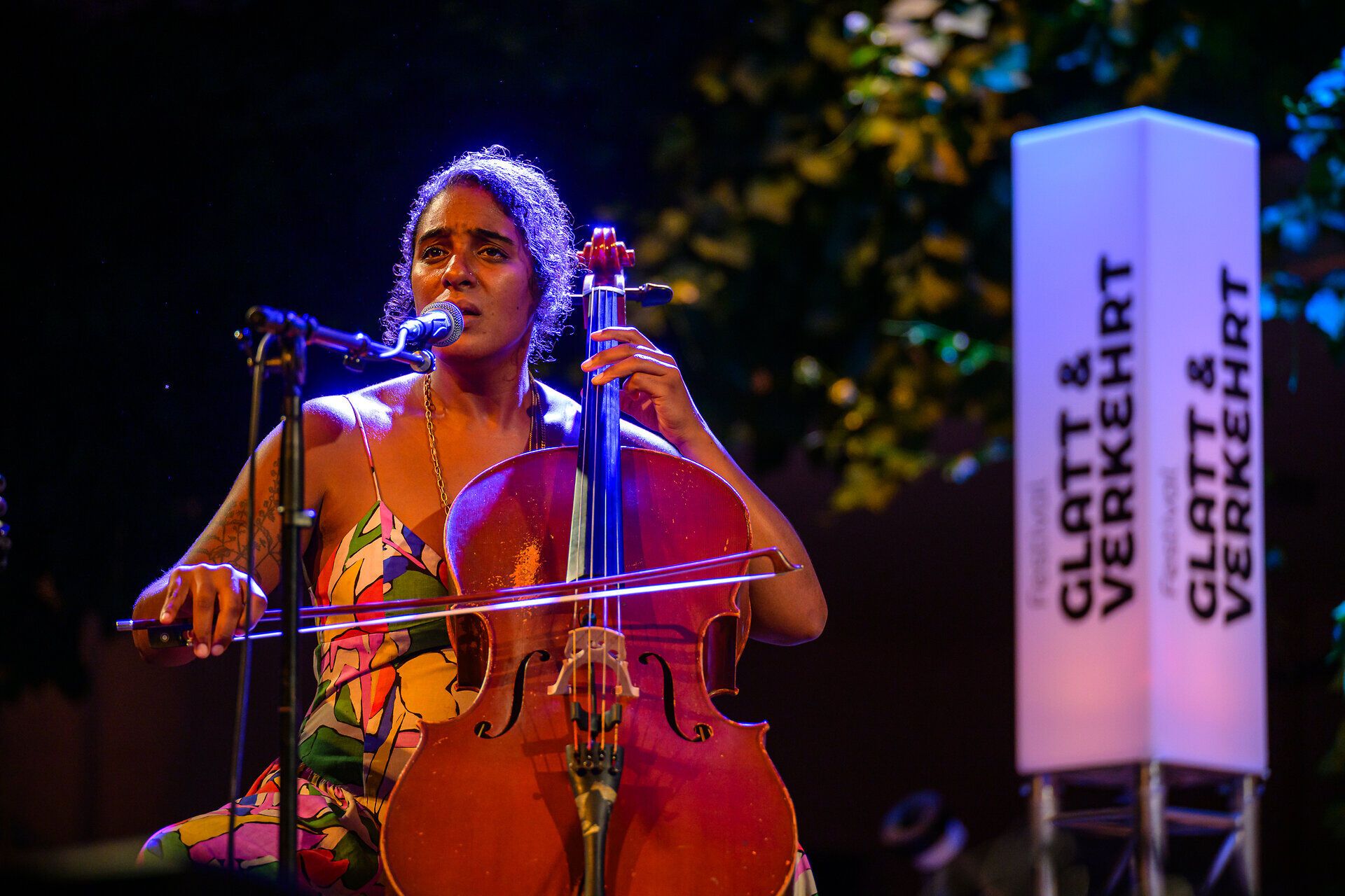 Cellistin und Sängerin steht auf der Bühne und spielt Cello vor Mikrofon beim Festival Glatt & Verkehrt, im Hintergrund beleuchtete Festival-Stele.
