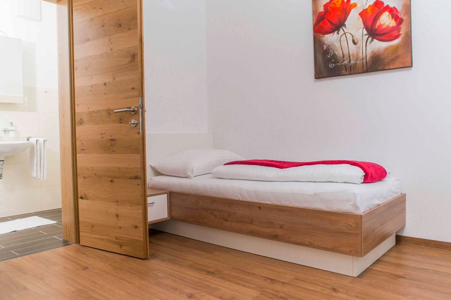 A simply furnished room with a single bed, wooden floor and a picture of red flowers on the wall.