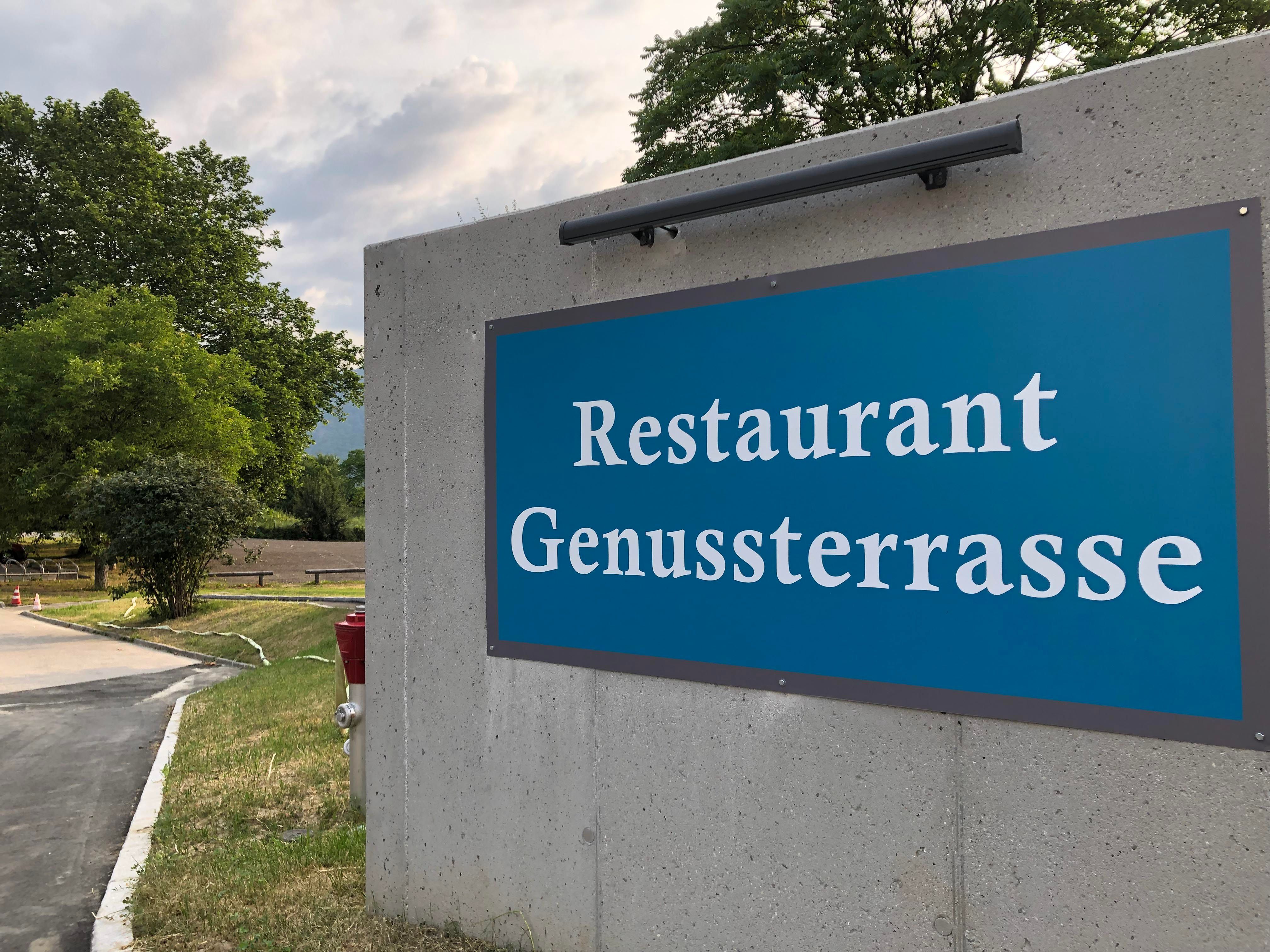 Sign for the restaurant 'Genussterrasse' on a concrete wall outside.