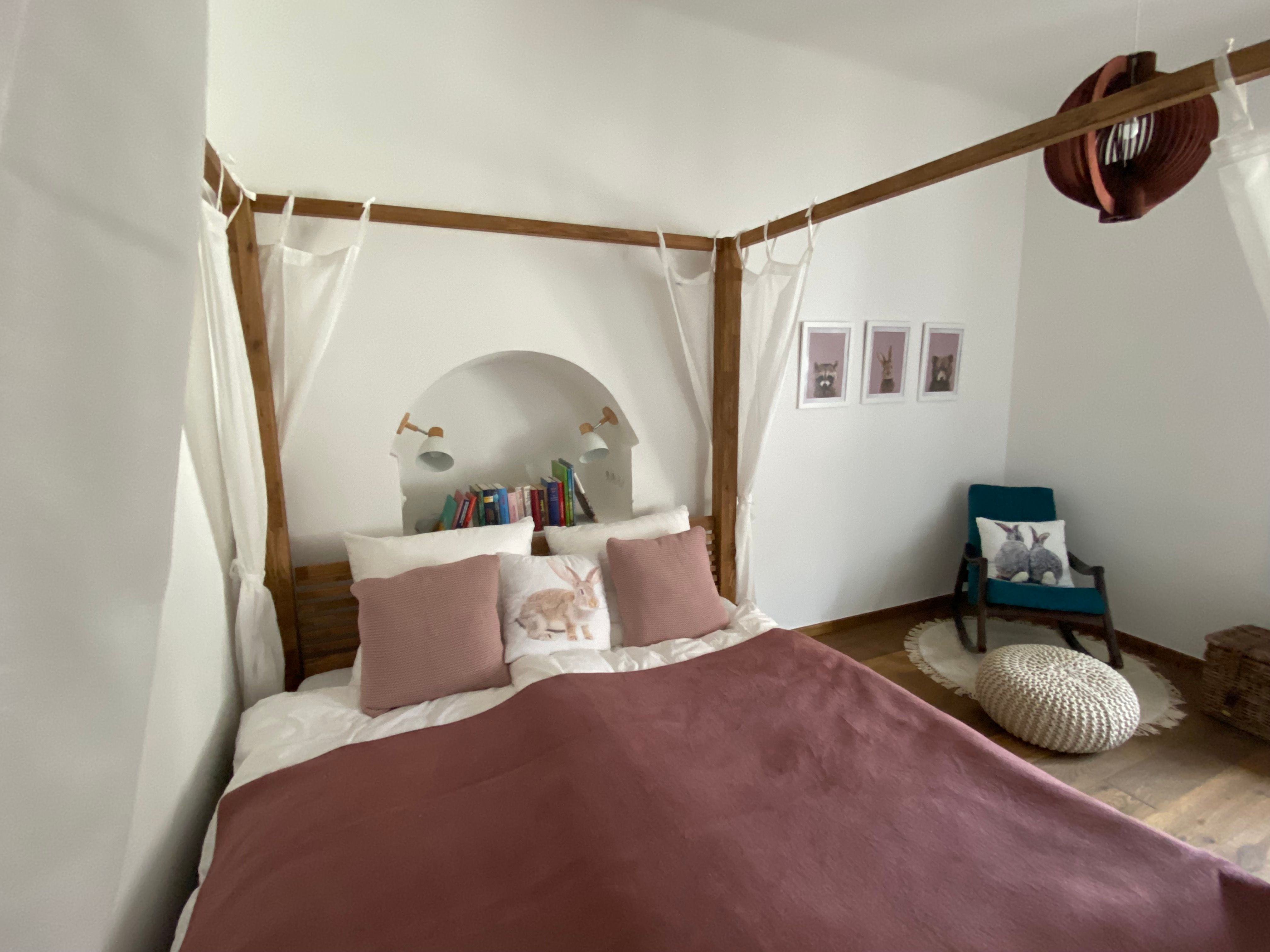 Cozy bedroom with four-poster bed, pillows, books and an armchair with rabbit cushions.