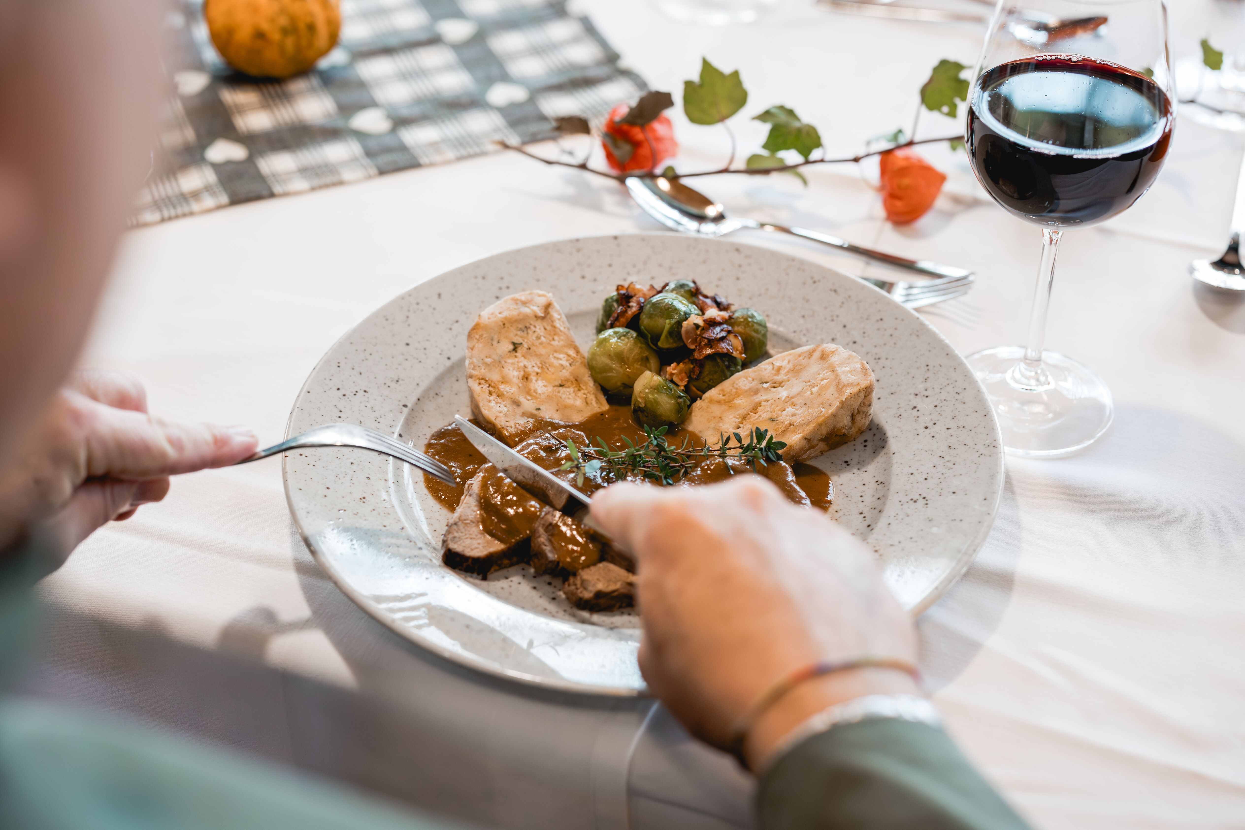 Person cuts meat on a plate with dumplings and Brussels sprouts, next to a glass of red wine.
