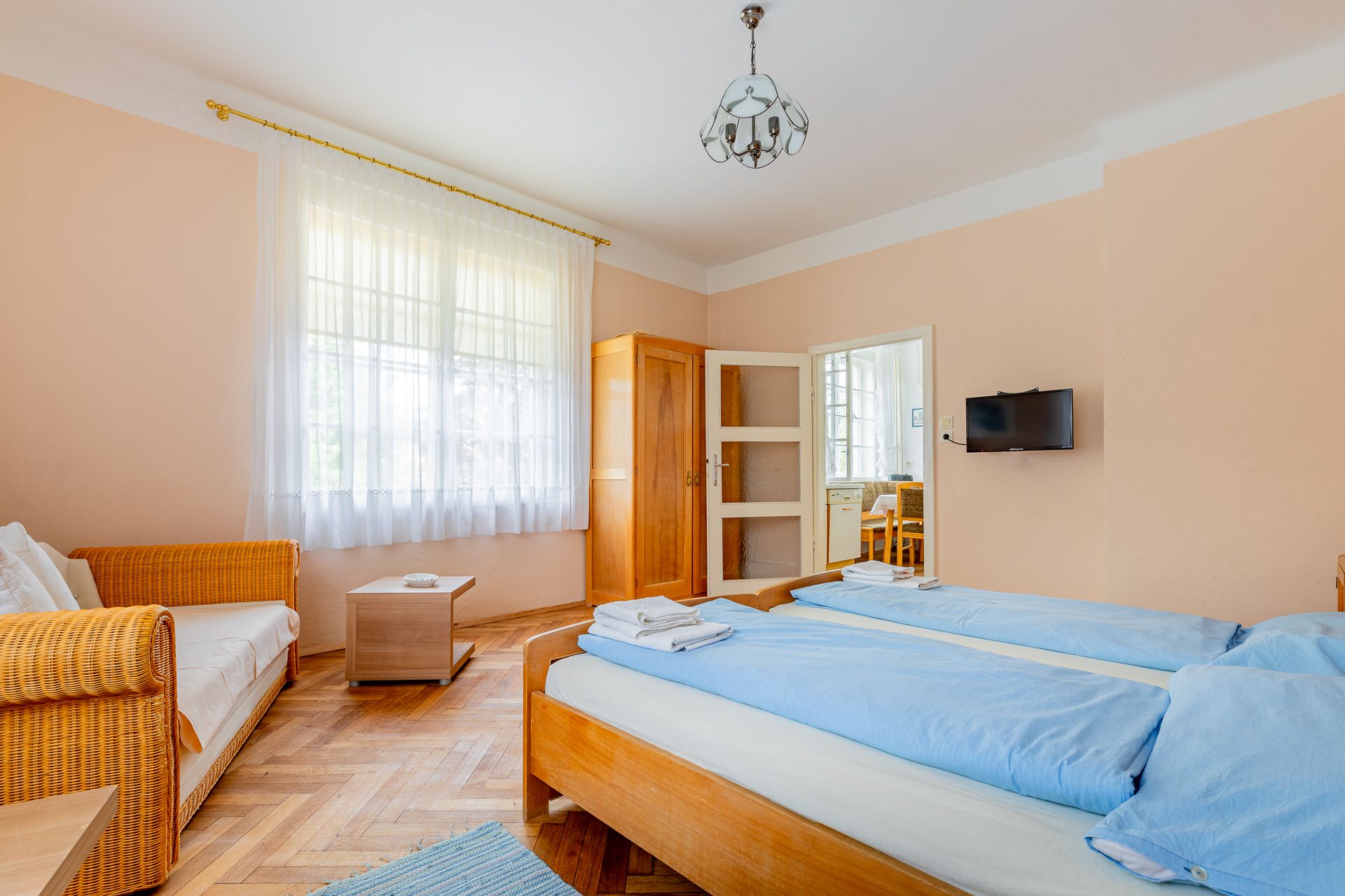 A cozy bedroom with two single beds, a sofa, a wardrobe and a small table. A window with curtains lets in natural light.