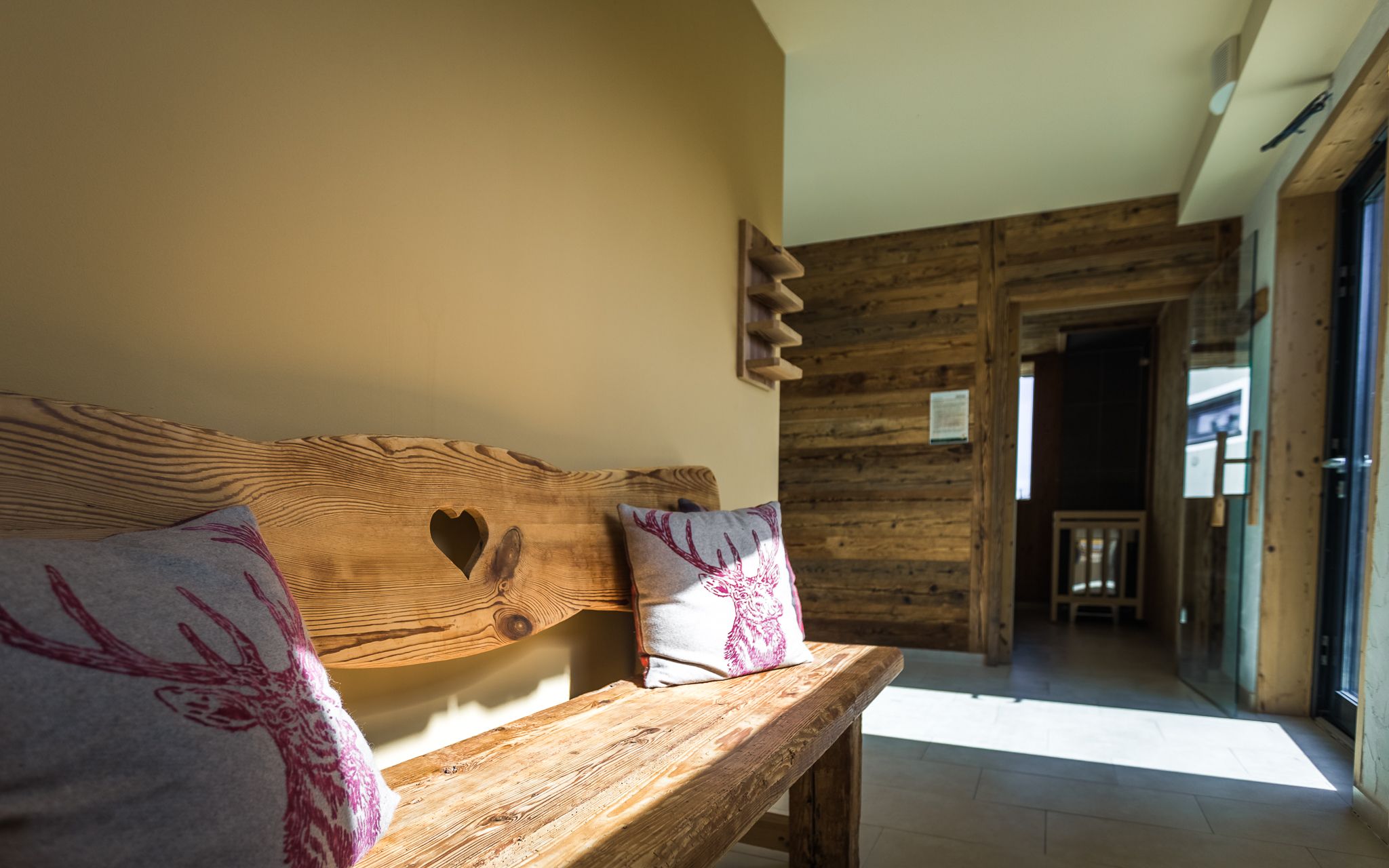 Rustic wooden bench with heart motif and deer cushions in a sunny room with wooden walls.