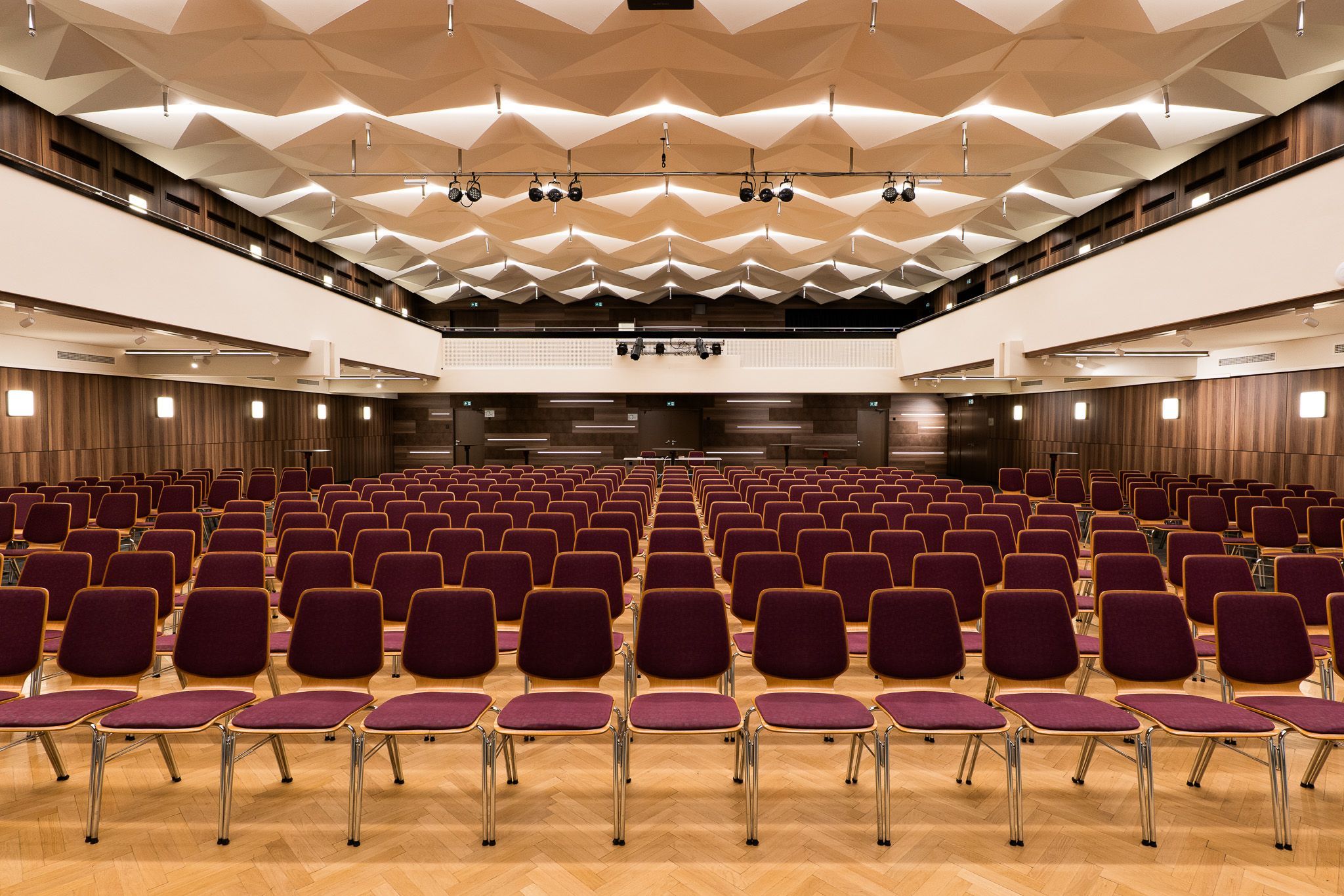 Empty event room with many rows of chairs and a geometric ceiling.