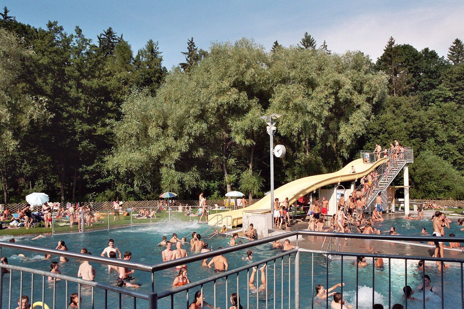 Outdoor pool with lots of people, slide and trees in the background.